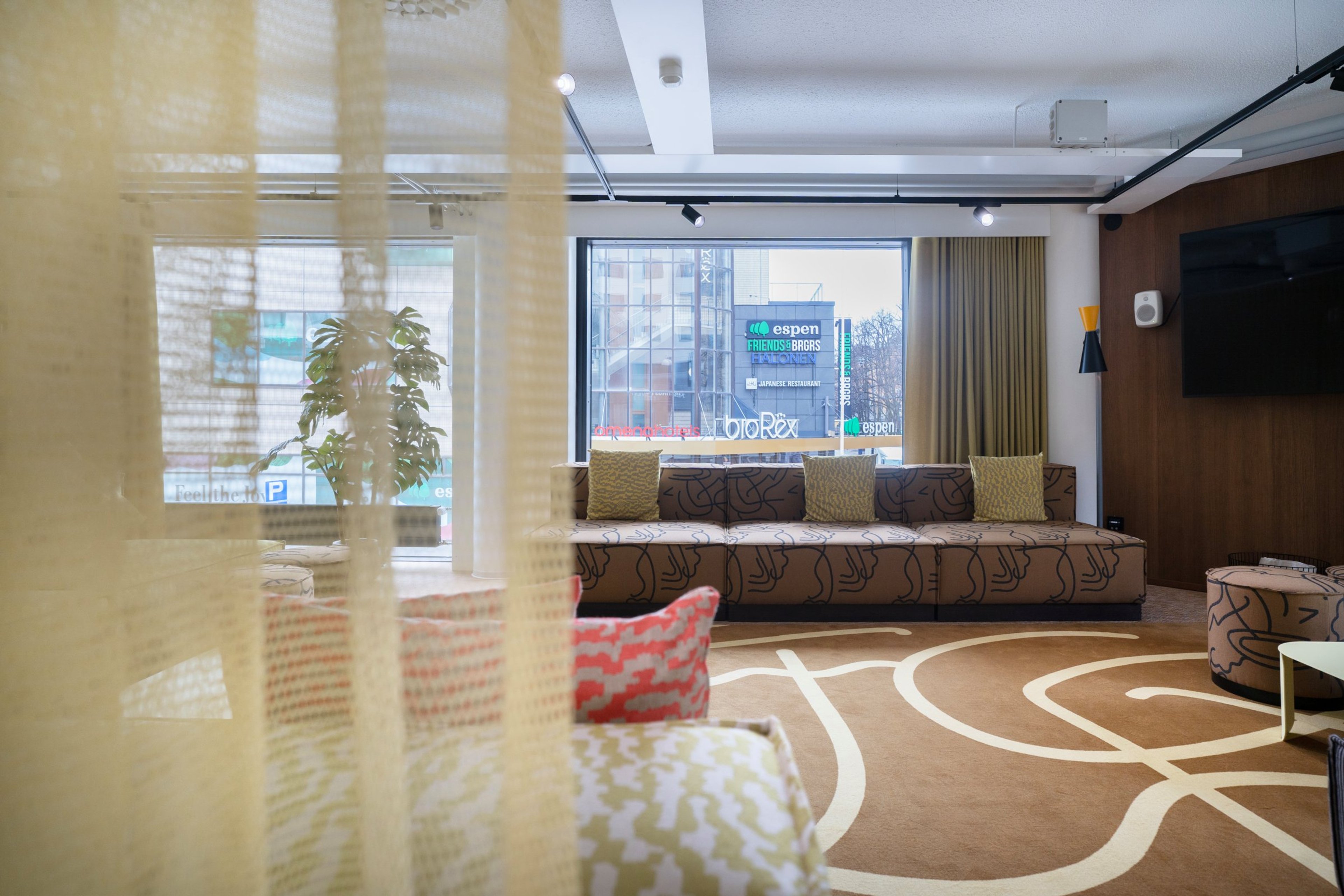 Moderni hotelliaula, jossa pitkä sohva ikkunan edessä, koristetyynyjä ja kuvioitu matto. Ikkunasta näkyy kaupunkinäkymä.