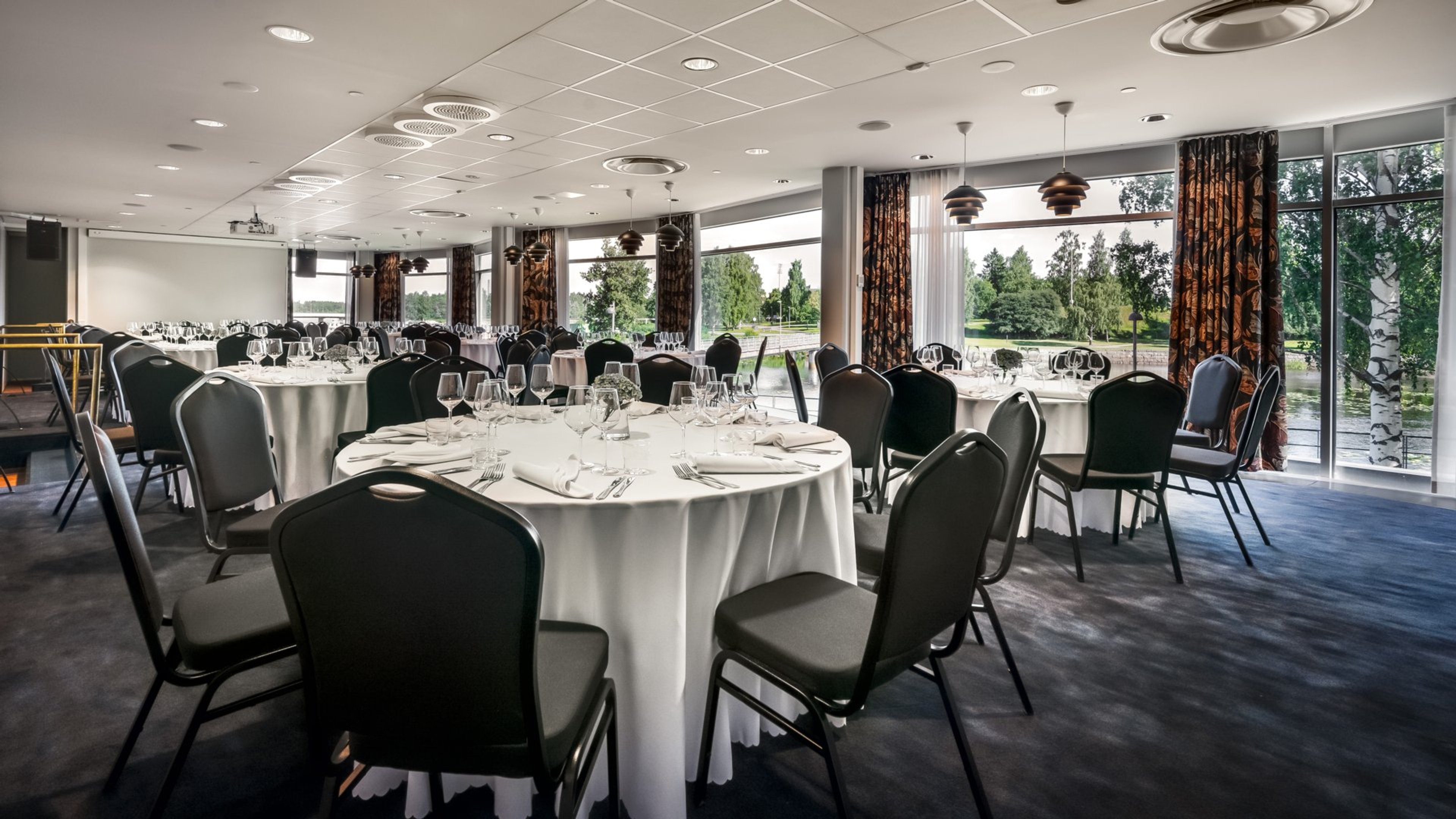 Juhlatila, jossa valkoisia pyöreitä pöytiä mustilla tuoleilla. Tila on valoisa suurten ikkunoiden ansiosta, joista näkyy puita ja vesialue.