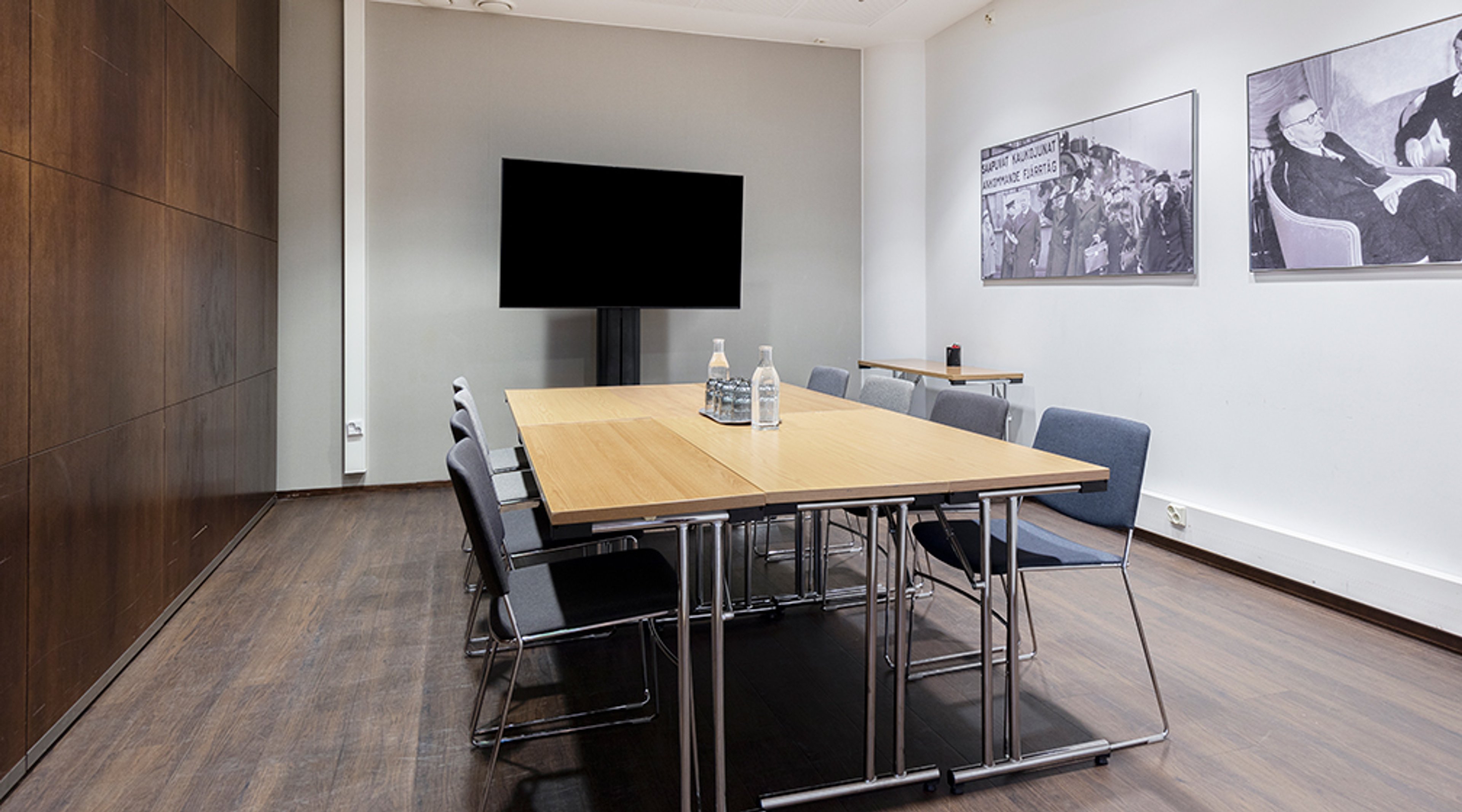 Moderni neuvotteluhuone puupöydällä, sinisillä tuoleilla, seinäasennettulla näytöllä ja historiallisilla valokuvilla. Tila on avara ja lattialla on tumma parketti.