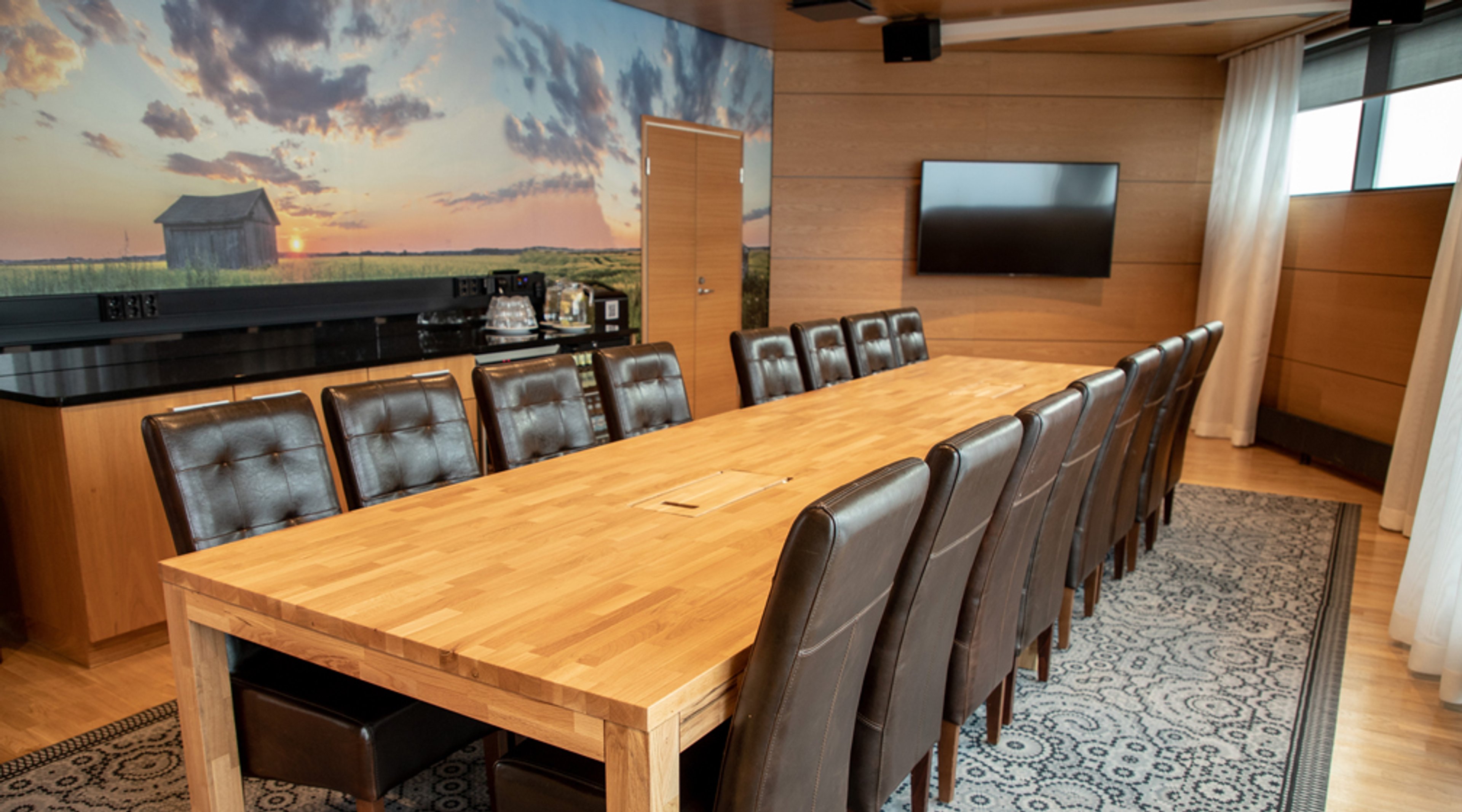 Neuvotteluhuone, jossa puinen pöytä, nahkatuolit, tv-näyttö ja panoraamaseinällä auringonlaskumaisema.