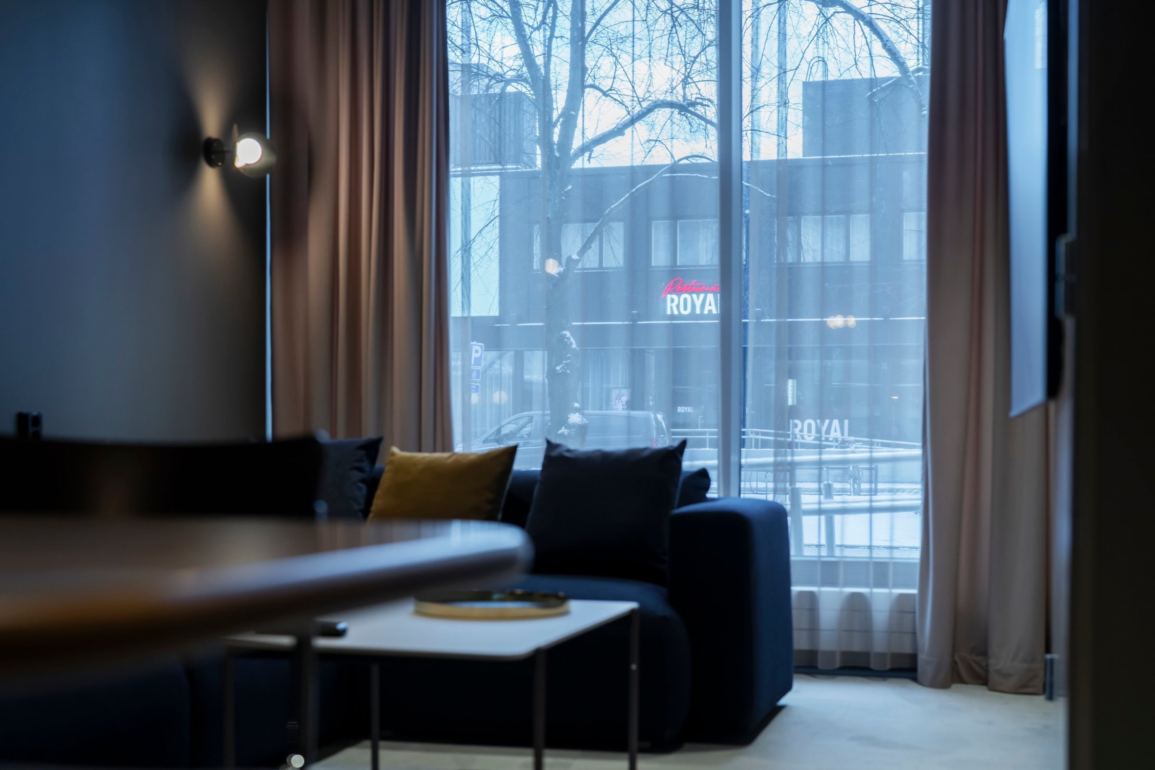 Hotellihuone, jossa sininen sohva keltaisella tyynyllä, suuret ikkunat ja näkymä toiseen rakennukseen. Huoneessa tasainen lattia ja hyvä valaistus.