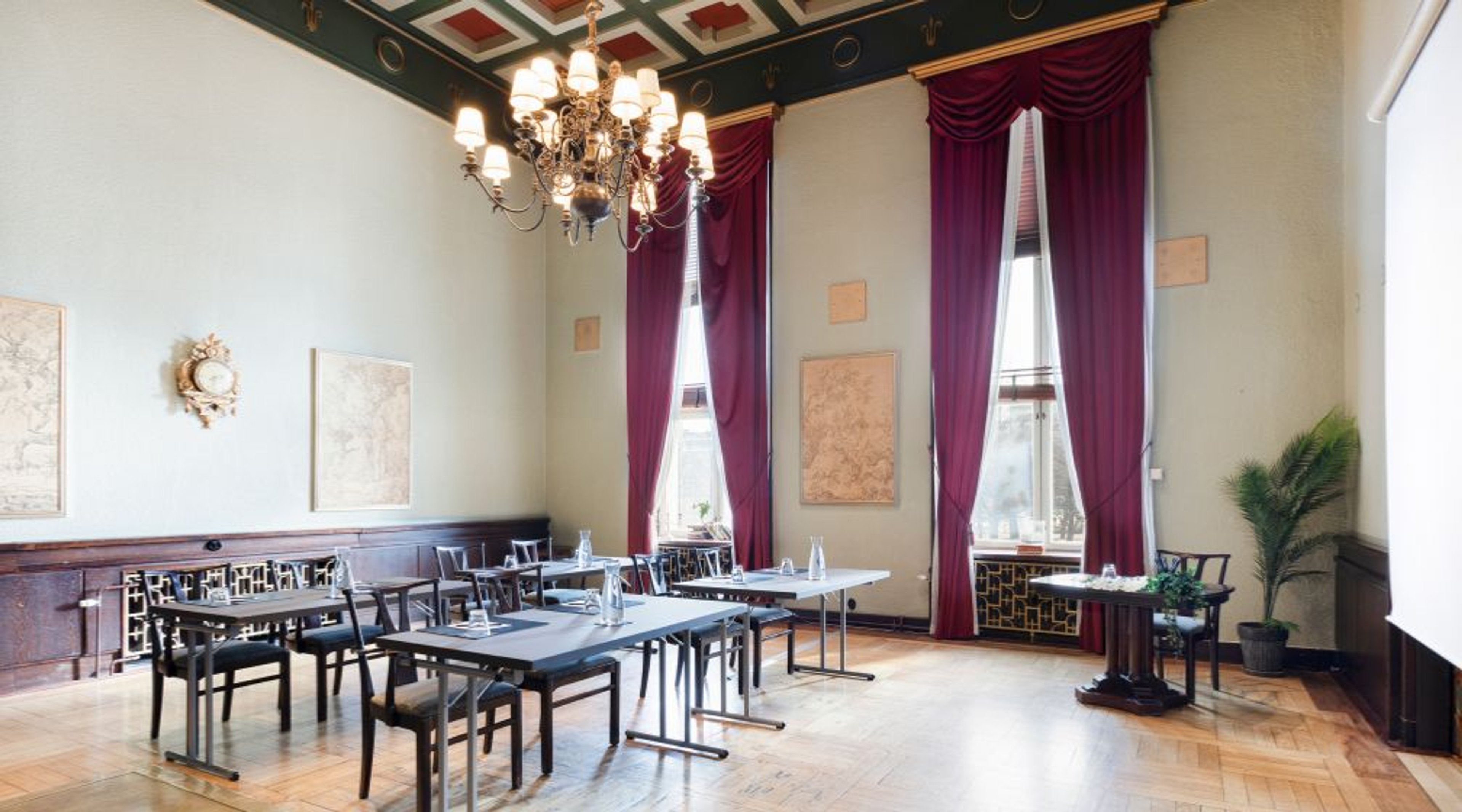 High-ceilinged conference room with tables and chairs, crystal chandelier, decorative ceiling painting, deep red velvet curtains and polished parquet flooring.