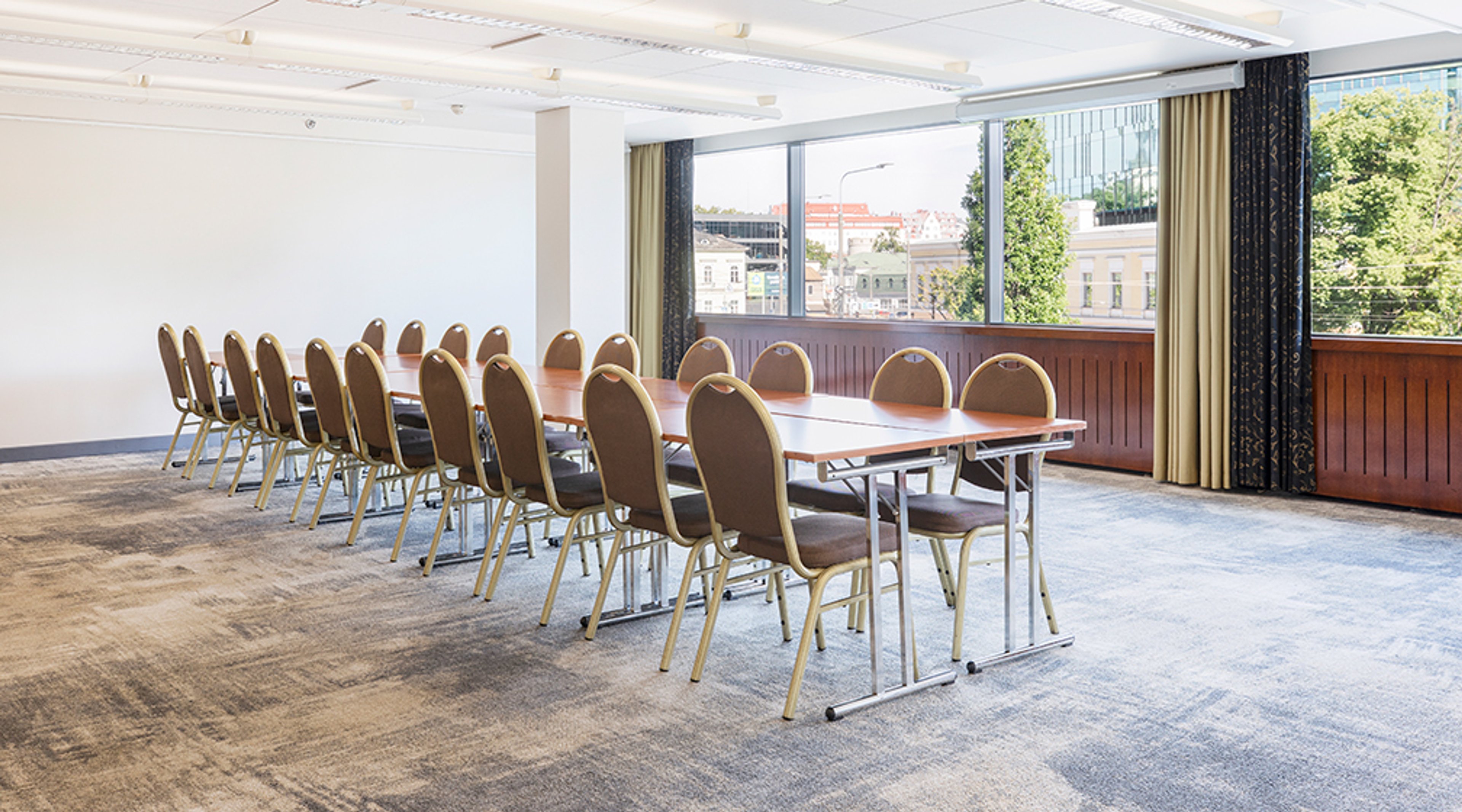 Conference room with a long table and brown upholstered chairs on both sides. Space is bright with large windows showing street view. Floor is level with low-pile carpet.