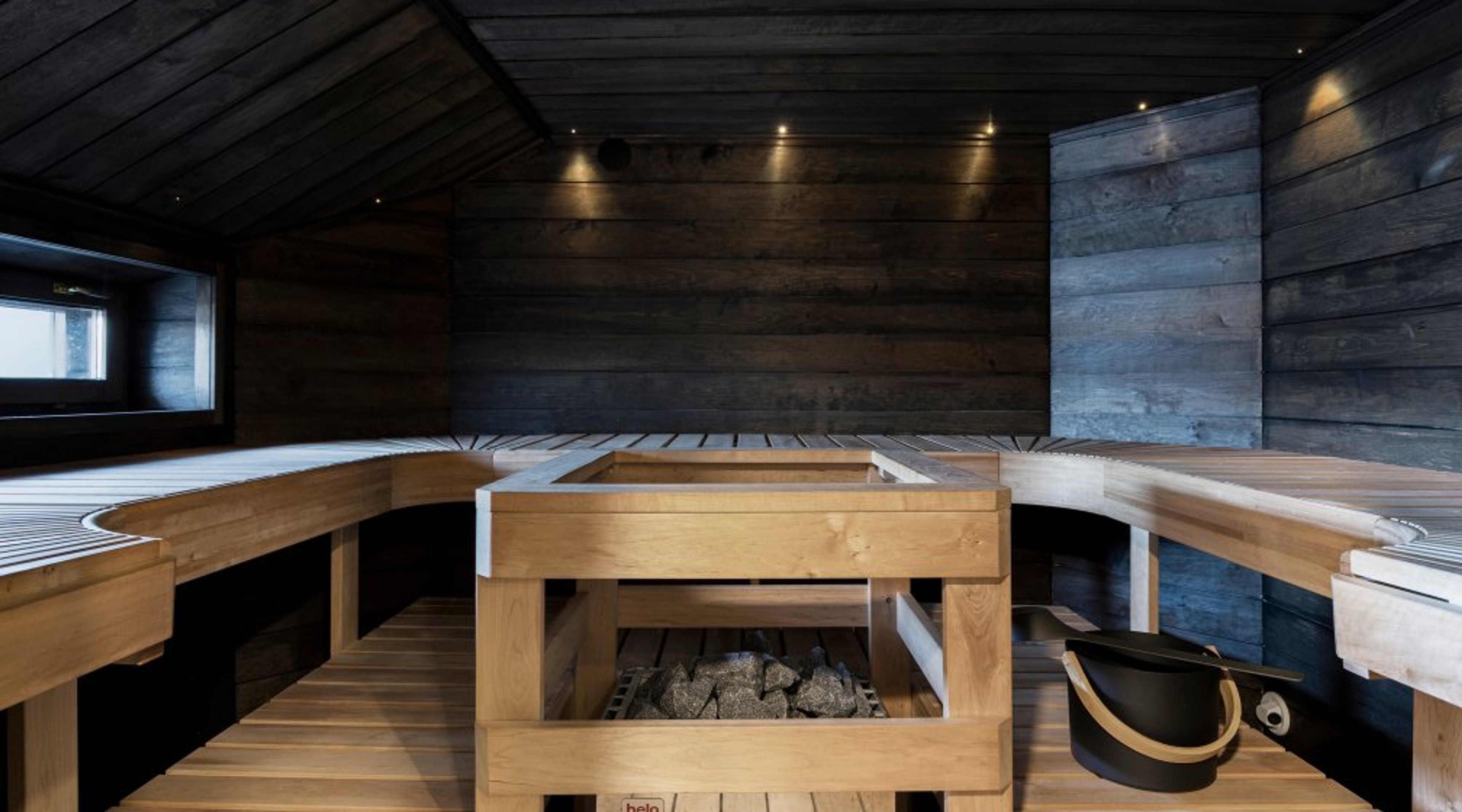 Modern sauna with dark wooden panels, light-colored seating benches and a heater in the center. Small window allows natural light in.