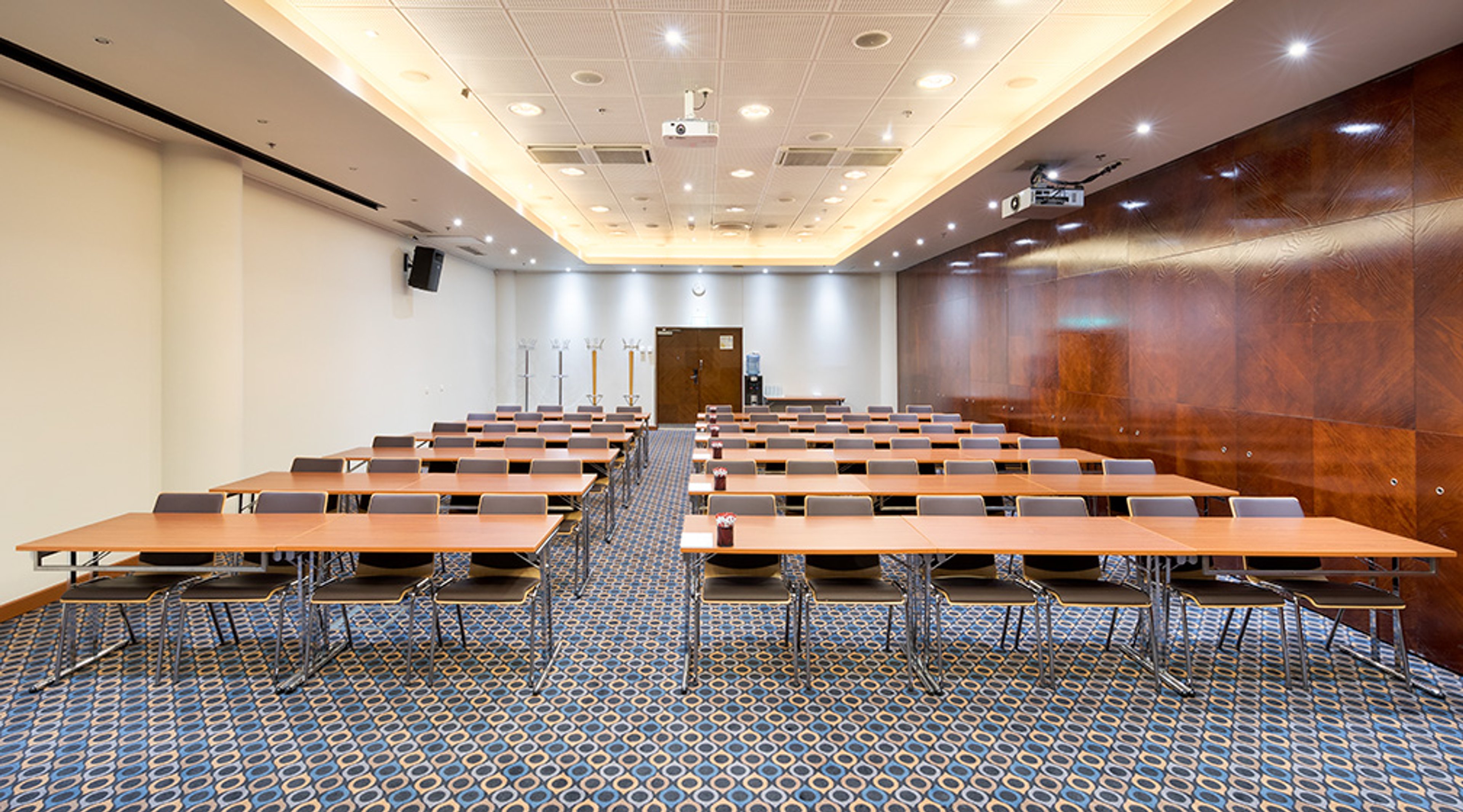 Kokoustila, jossa on riveihin järjestetyt pöydät tuoleineen, ruskeat puupaneelit seinällä, sininen kuvioitu kokolattiamatto ja kattoon upotetut valot.