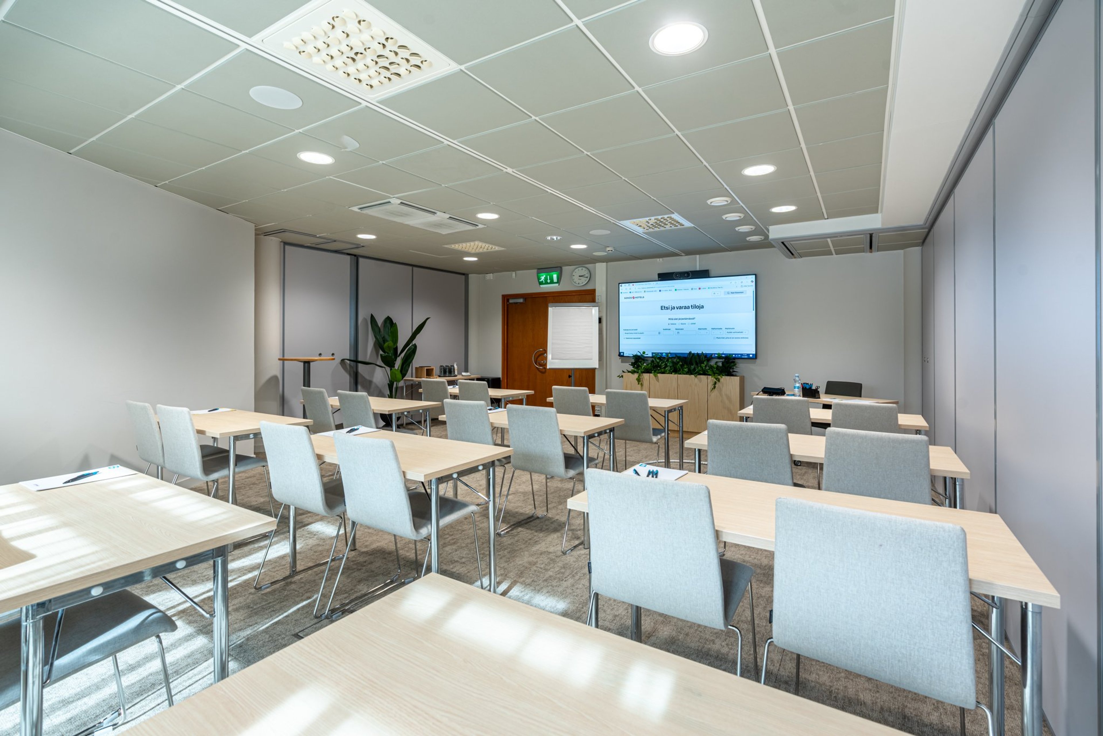 Moderni kokoustila vaaleilla pöydillä ja tuoleilla. Huoneessa on suurinäyttö, tasainen lattiapinta ja hyvä valaistus.