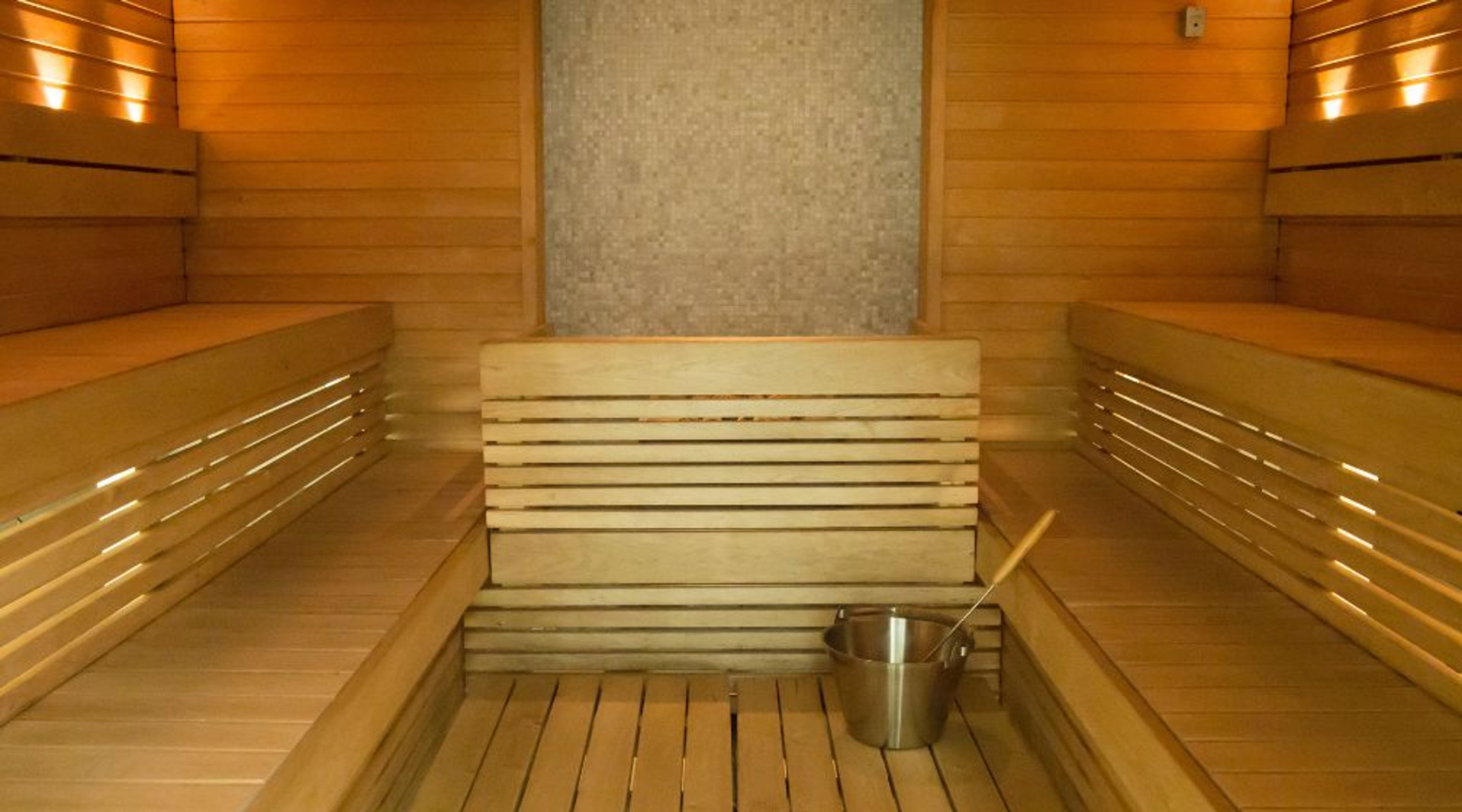 Moderni sauna puisilla lauteilla, taustan mosaiikkiseinä ja metallikauha lattialla.