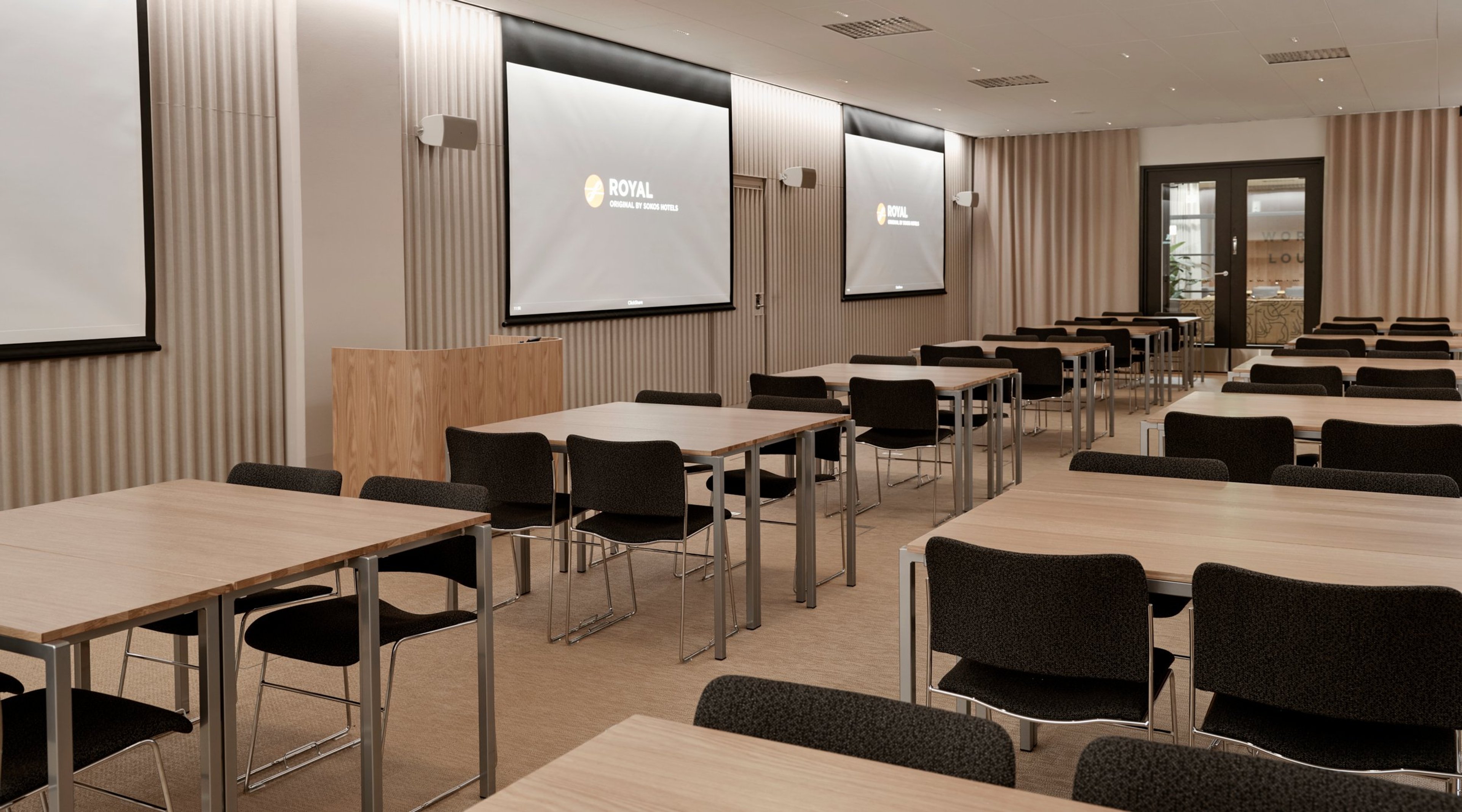 Moderni konferenssihuone puisilla pöydillä ja mustilla tuoleilla. Kolme valkokangasta edessä, tasainen lattia ja hyvä valaistus.