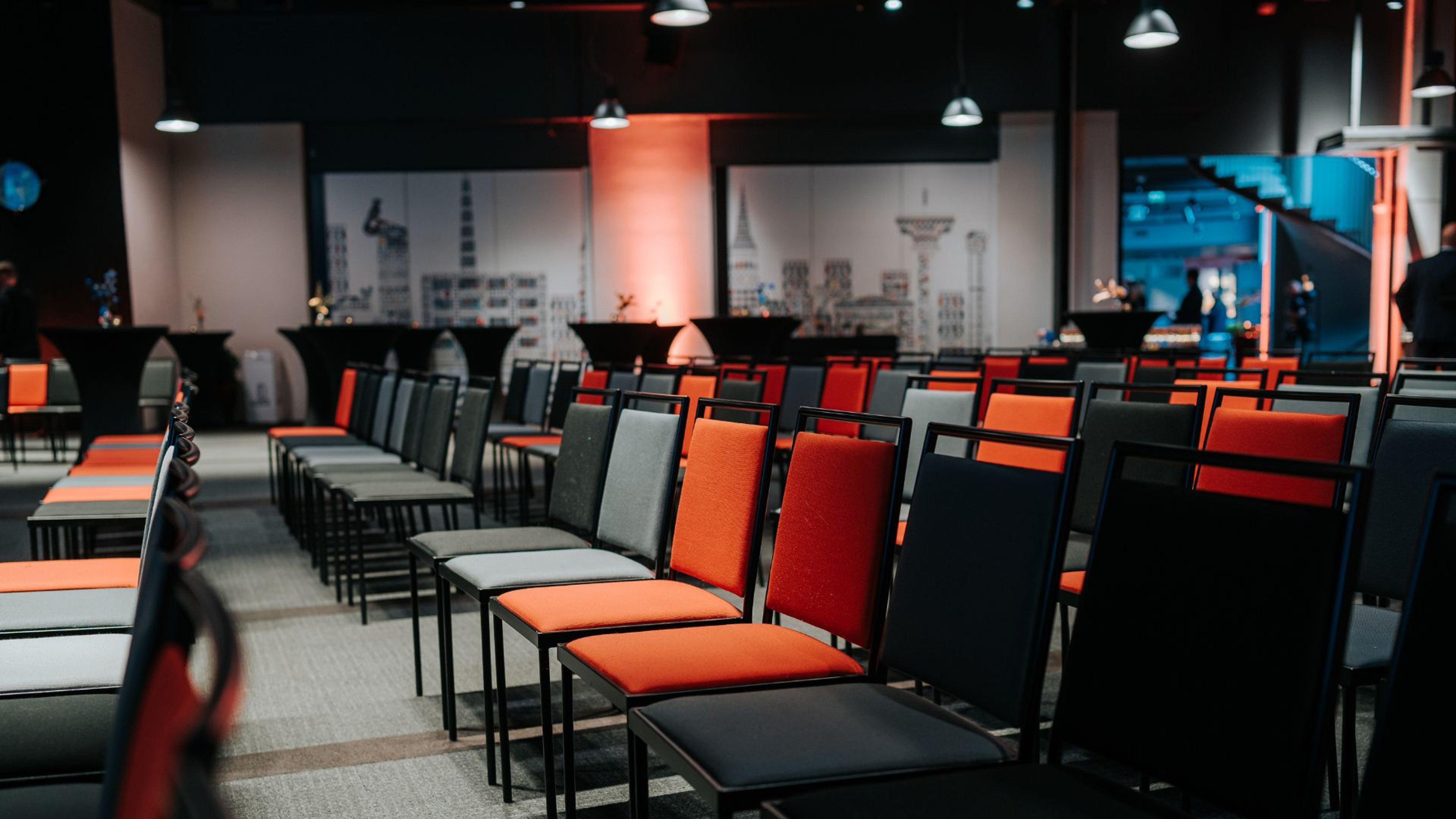 Konferenssitila, jossa on mustia, punaisia ja harmaita tuoleja riveittäin. Taustalla on kaupunkimaisemaa kuvaavat seinäpaneelit ja korkeakattoisessa tilassa on riippuvalaisimia.