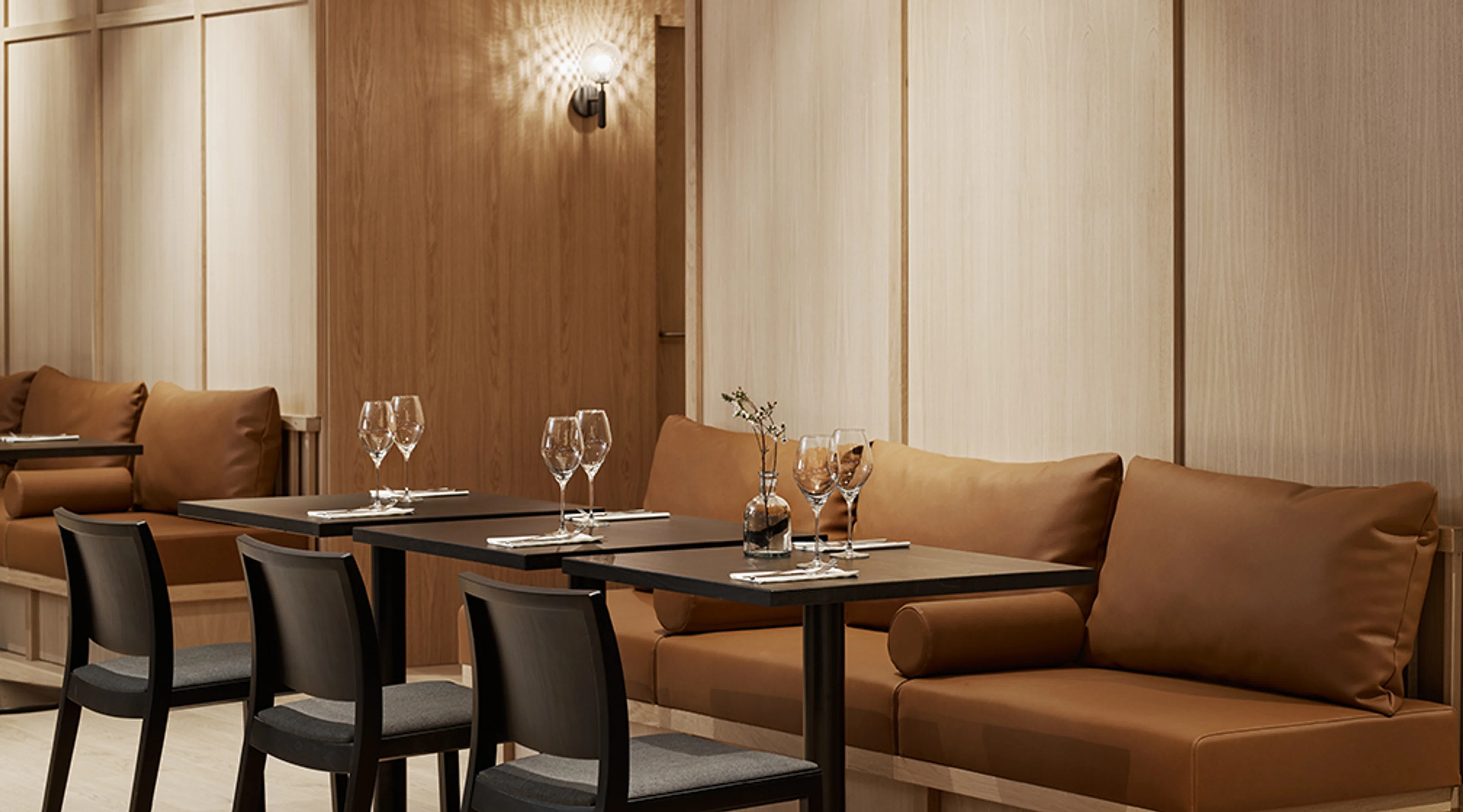Moderni ravintola sisustettuna puupaneloinnilla, ruskeilla sohvilla ja mustilla pöydillä. Pöydillä viinilaseja ja kattauksia.