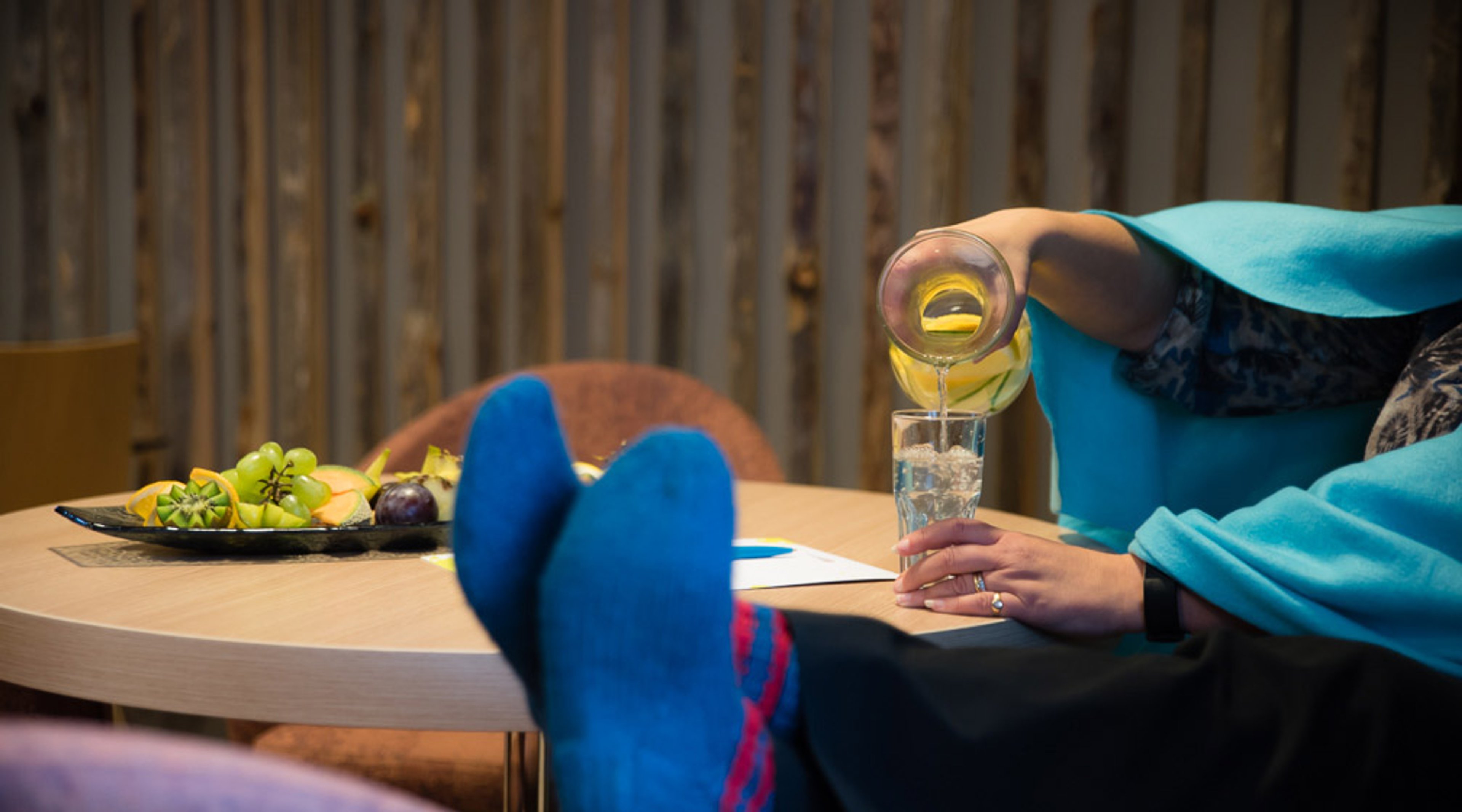 Henkilö kaataa keltaista juomaa jäillä täytettyyn lasiin. Pöydällä on hedelmälautanen ja etualalla näkyy siniset sukat.