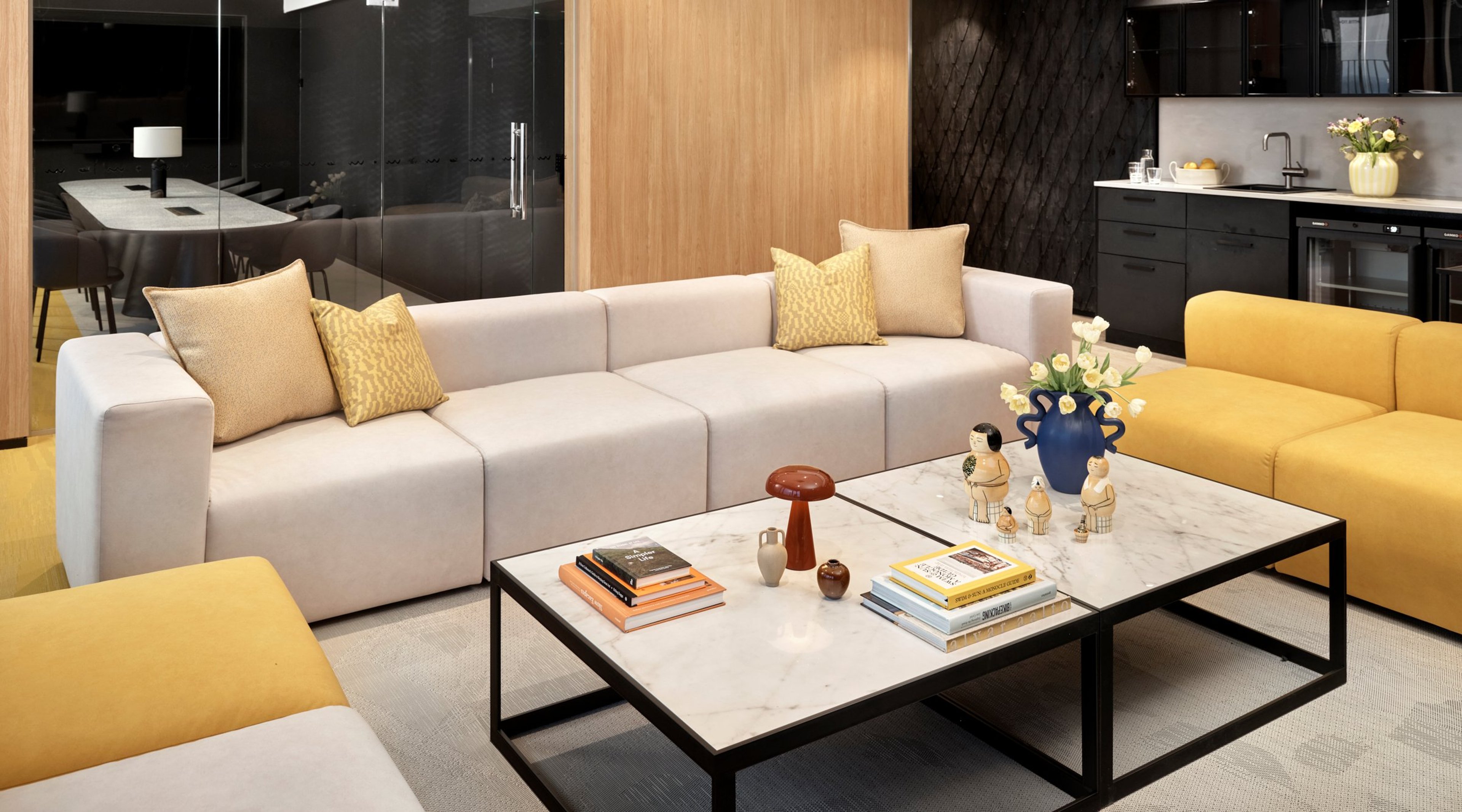Moderni olohuone, jossa valkoinen sohva keltaisilla tyynyillä, keltainen nojatuoli ja marmoripintainen sohvapöytä. Taustalla lasiseinä ja pieni keittiö.