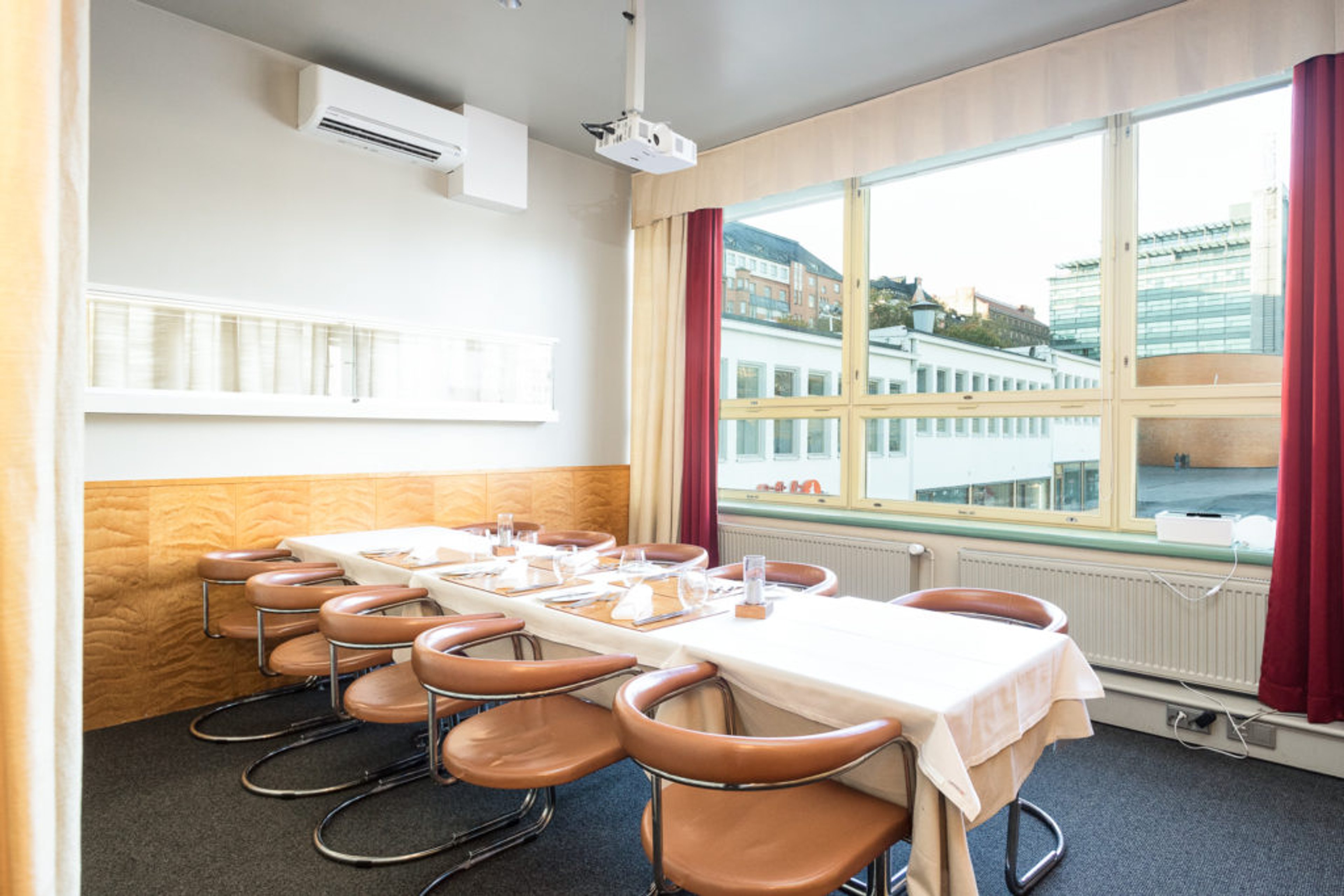 Konferensrum med ett långt bord täckt med en ljus duk. Läderfåtöljer runt bordet. Luftkonditionering och projektor på väggen. Stadsutsikt genom fönstret.