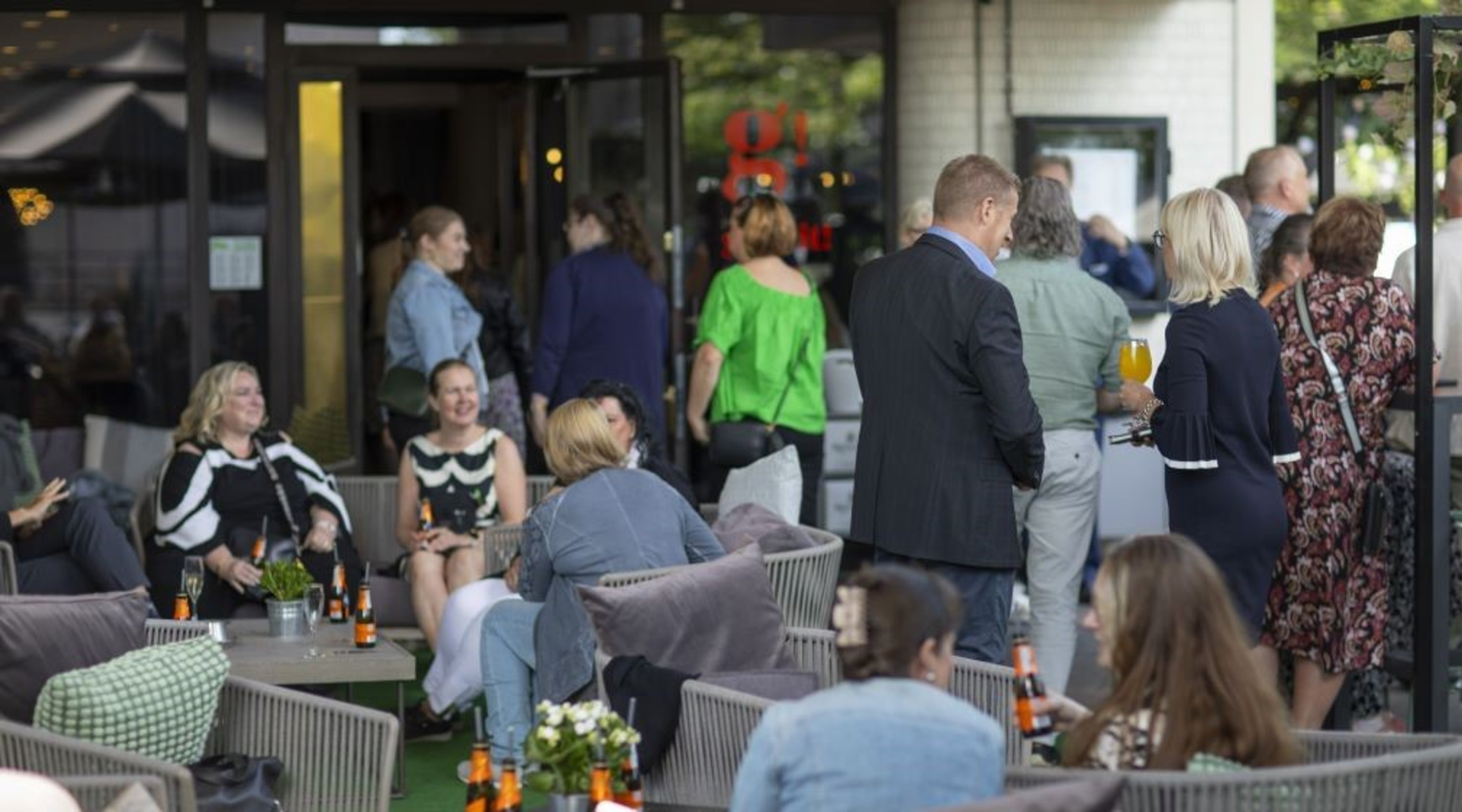 Ihmisiä viihtyisällä terassi- tai ravintola-alueella, jossa ryhmä istuu vaaleissa rottinkituoleissa nauttien juomia ja seurustelua.