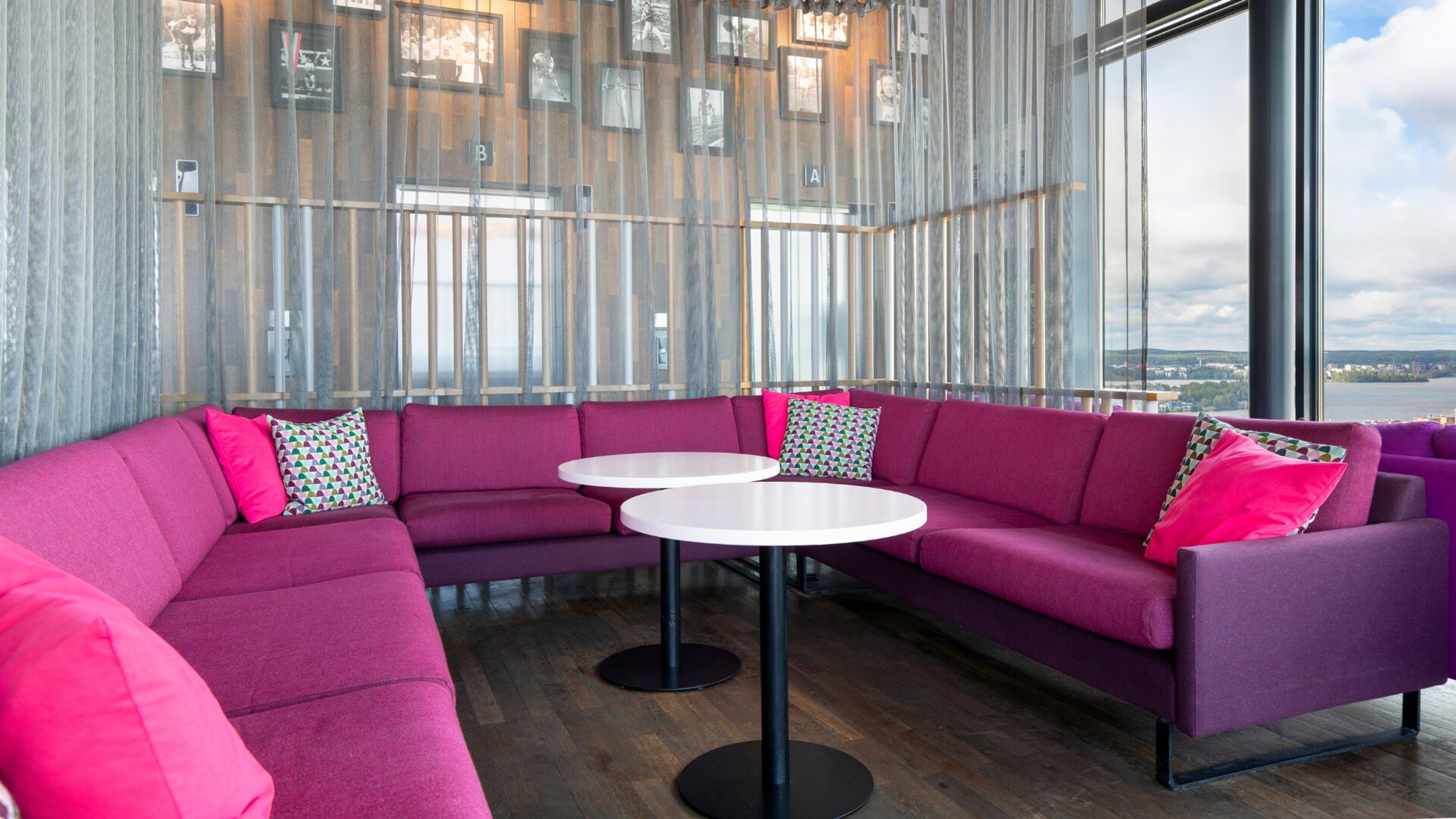 Pinkki kulmasohva värikkäillä tyynyillä ja kaksi pyöreää pöytää oleskelutilassa. Isot ikkunat tarjoavat näkymän järvelle ja kaupunkiin.