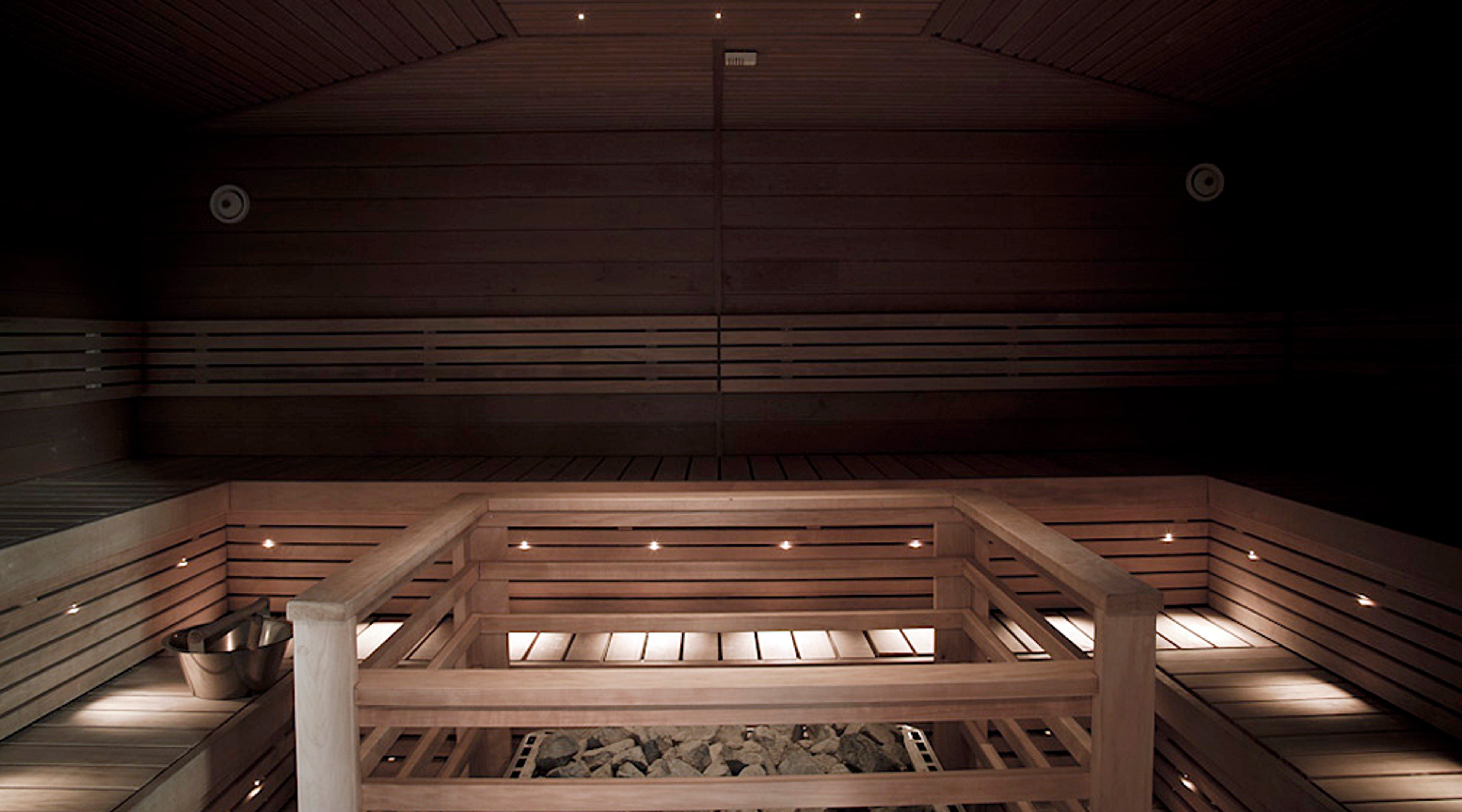 Hämärä puusauna sisältäpäin kuvattuna. Etualalla näkyy puiset lauteet ja kiuas, jossa kiuaskivet. Minimalistiset valot tuovat tunnelmaa.