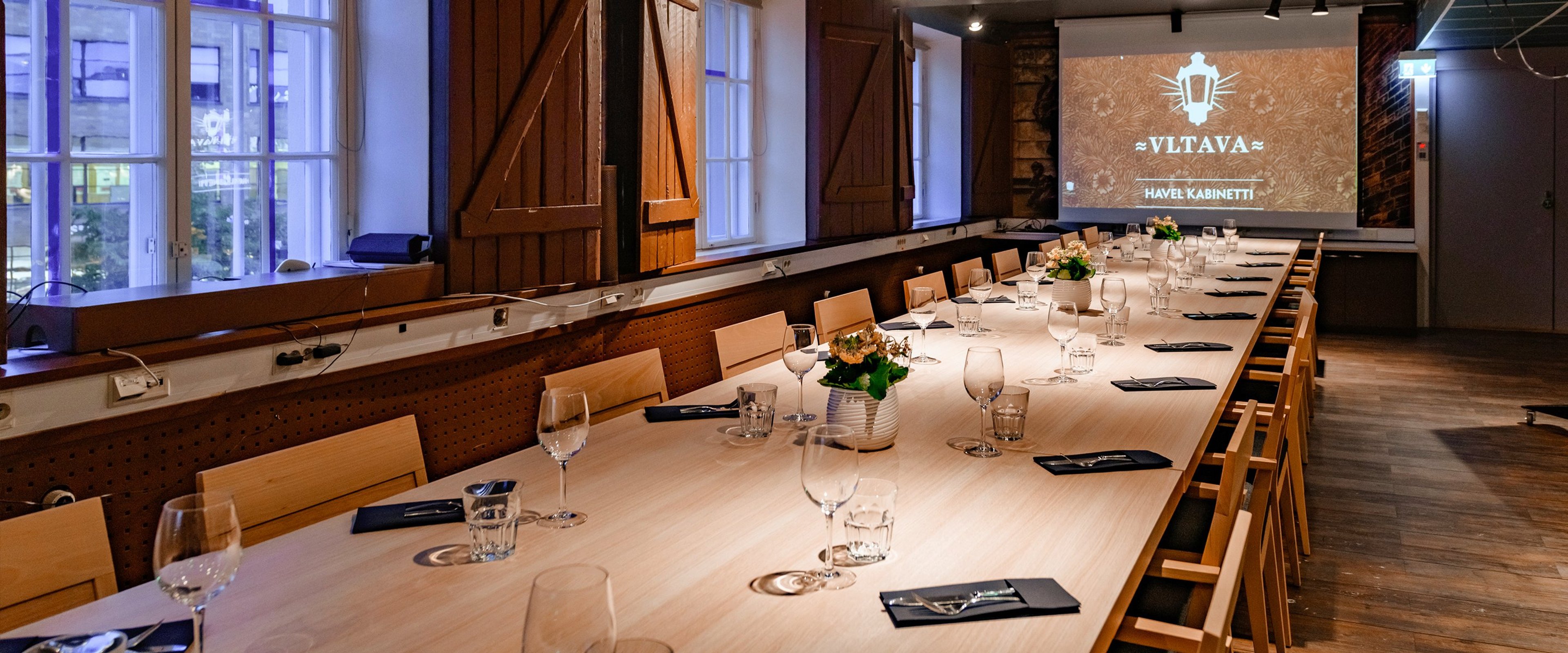 Pitkä pöytä kokoustilassa puisilla tuoleilla, laseilla ja ruokaliinoilla katettuna. Seinällä projektori näyttää Vltava-logoa.