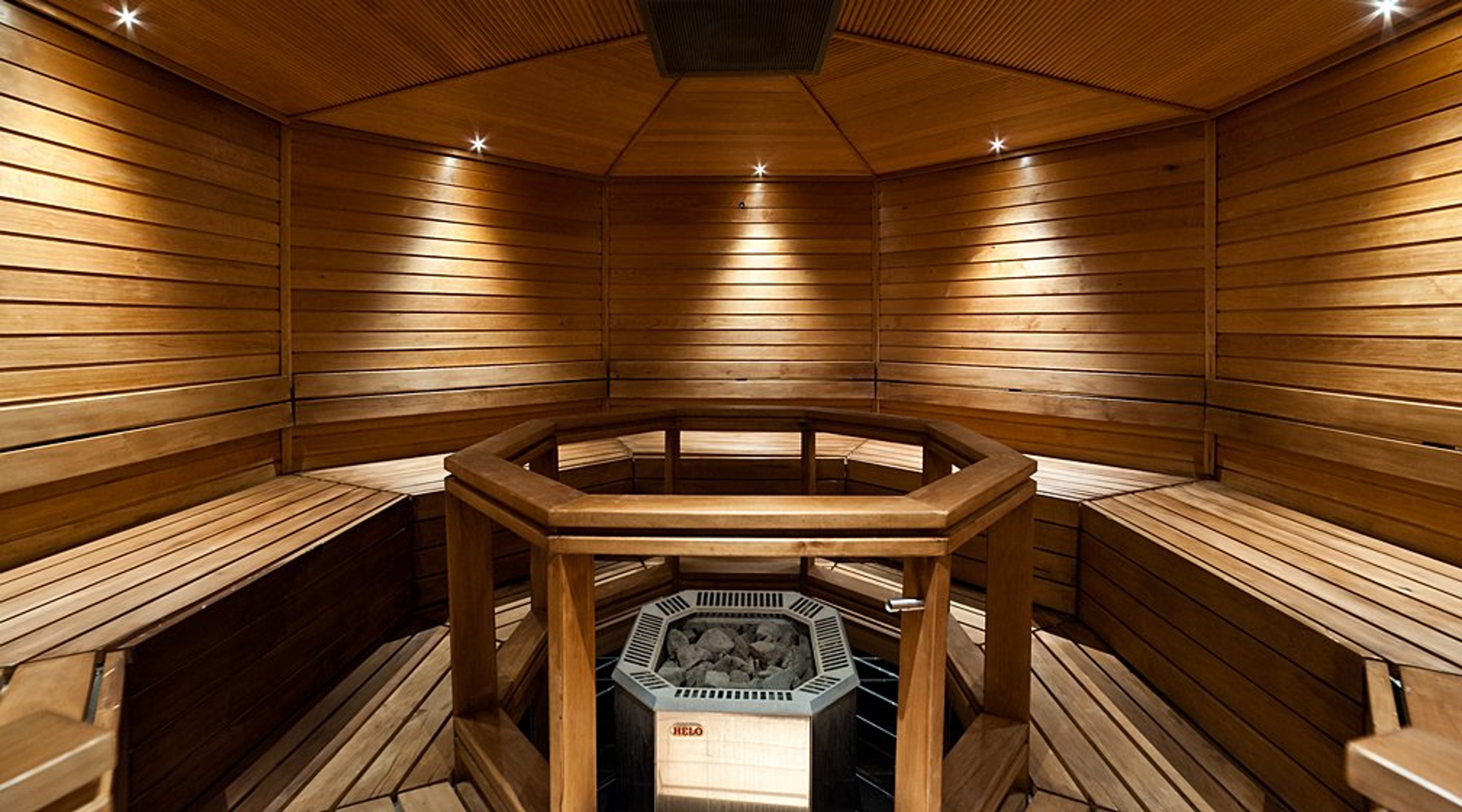 Kuusikulmainen sauna, jossa puiset lauteet reunustavat seinustoilla keskellä olevaa kiuasta. Puupaneloidut seinät ja kattovalaistus.