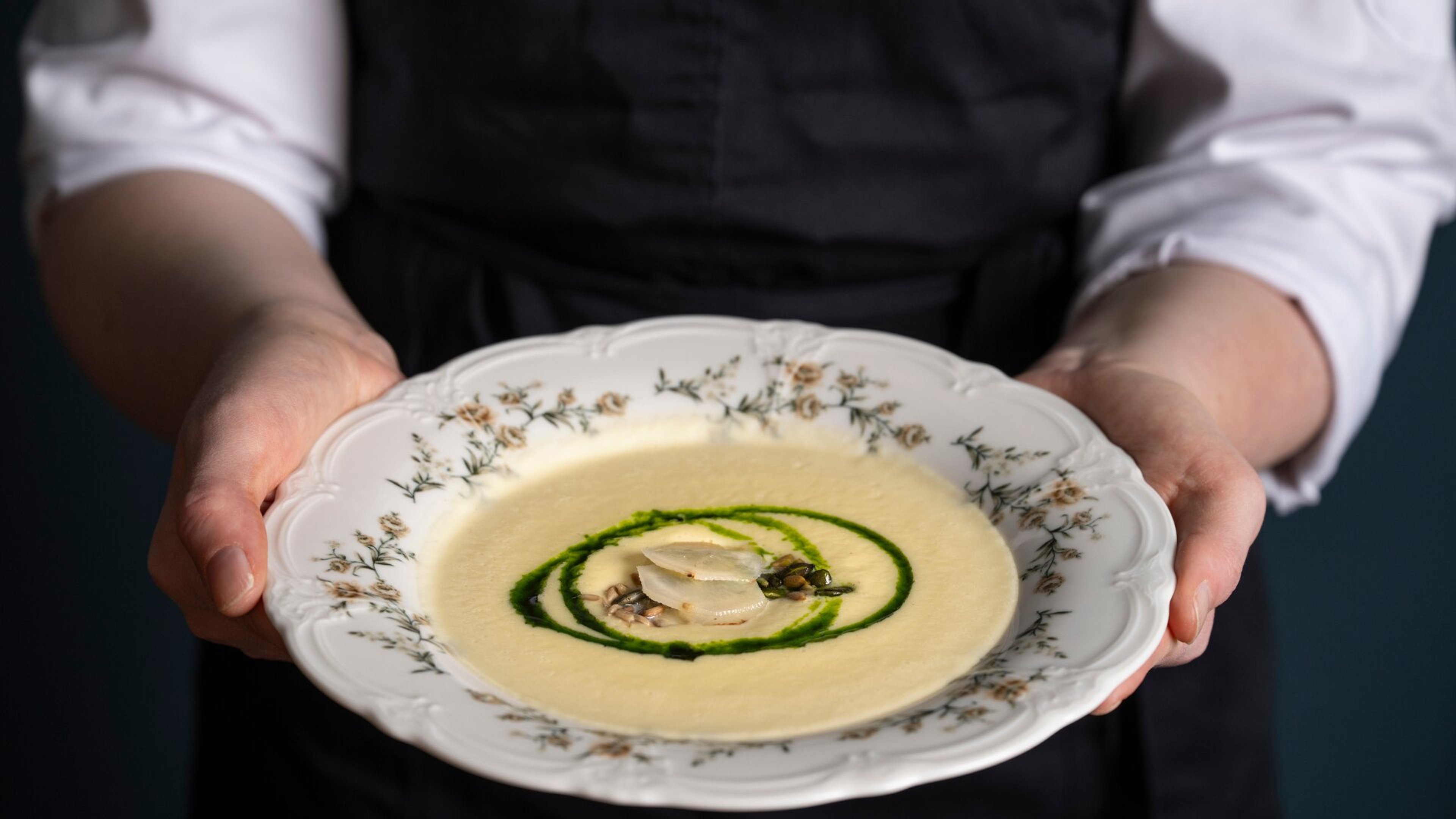 Henkilö mustassa esiliinassa tarjoilee koristeellisessa posliinilautasessa vaaleaa keittoa, jossa vihreä spiraalikuvio ja merellisiä lisukkeita keskellä.