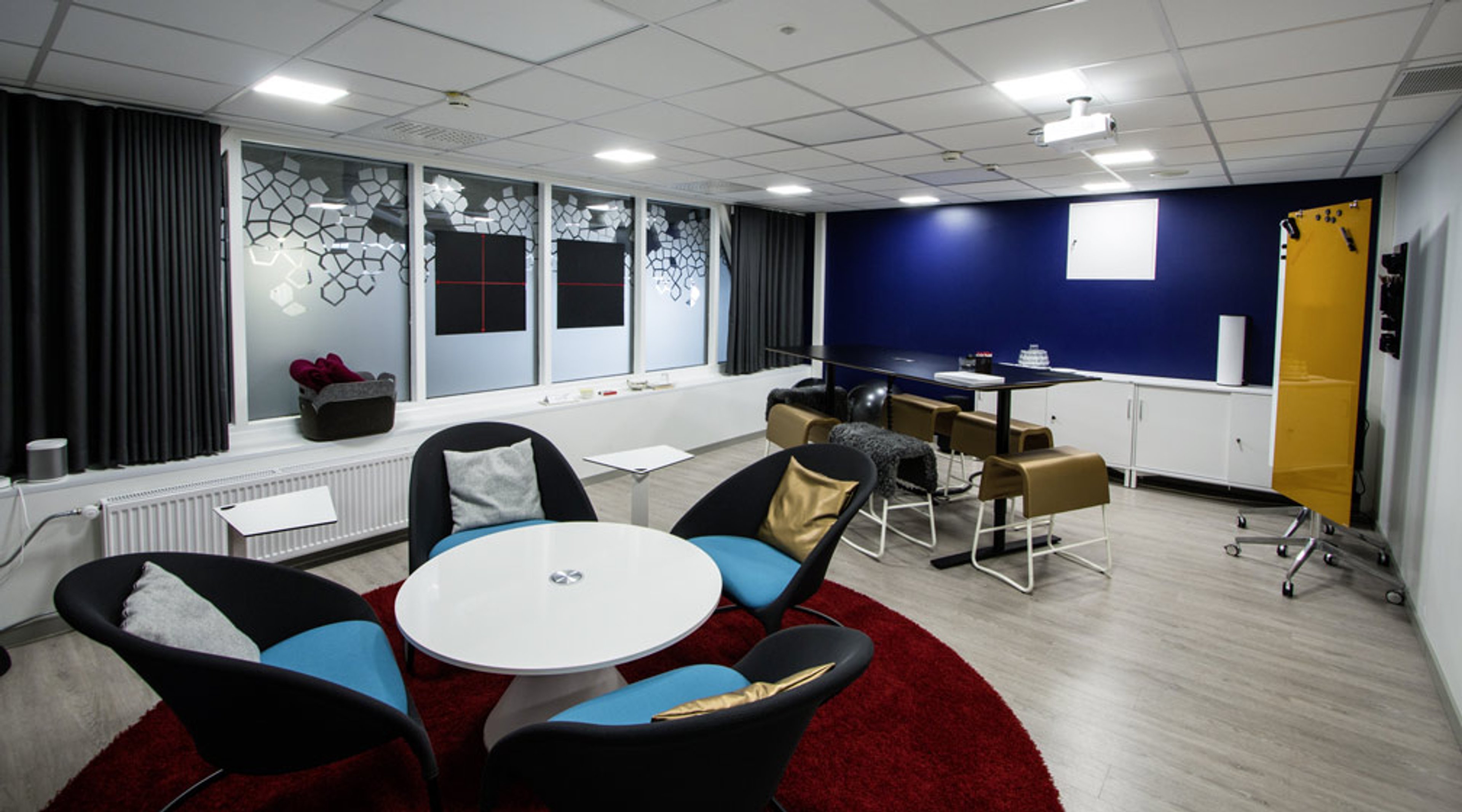 Moderni kokoontumistila, jossa pyöreä pöytä sinisillä tuoleilla punaisella matolla ja takaosassa pitempi pöytä ruskeilla tuoleilla. Sininen seinä ja projektori.