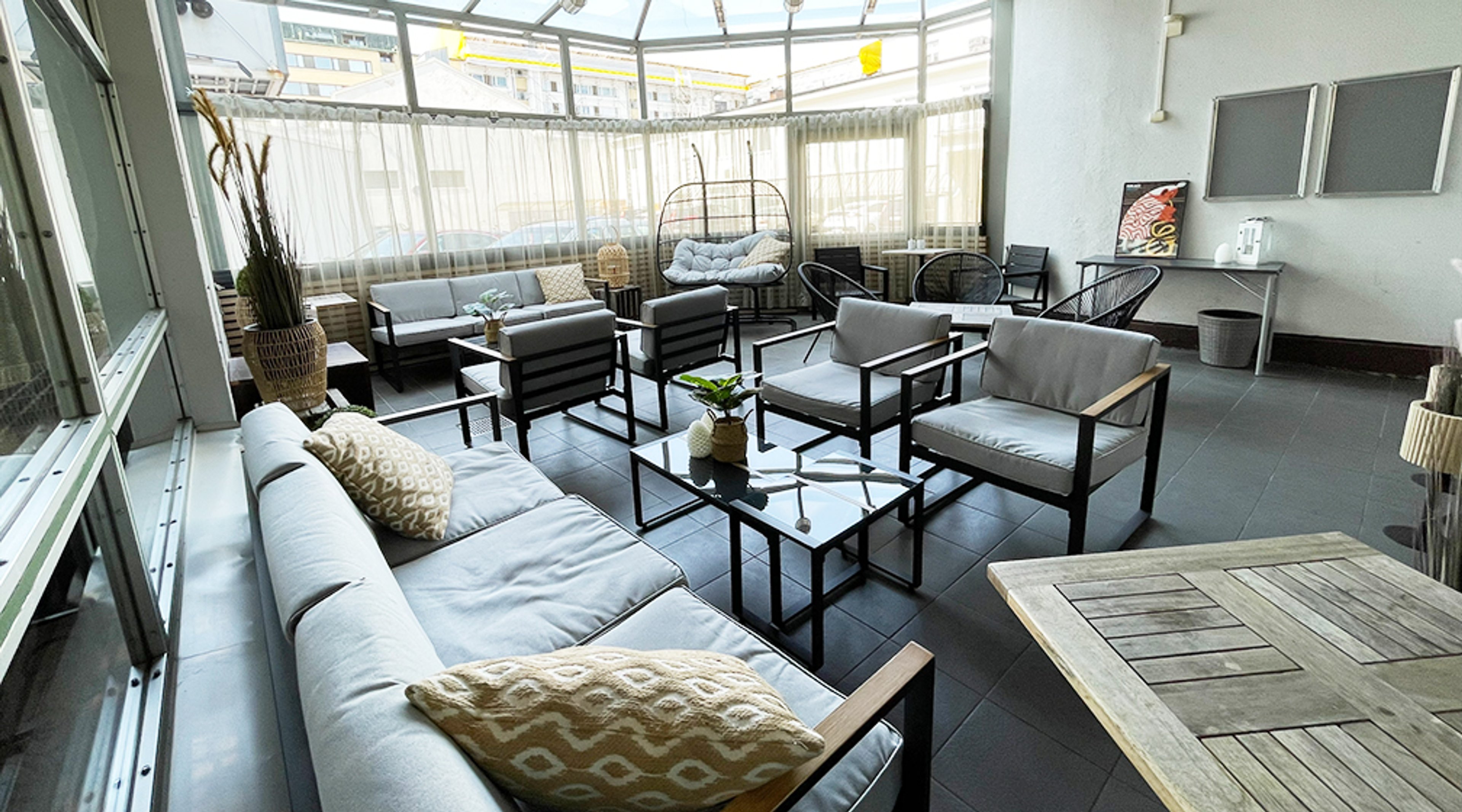 Valoisa lasitettu terassi, jossa harmaita sohvia ja nojatuoleja, keskellä lasinen sohvapöytä. Oikealla riippuva tuoli ja pieni pöytä.