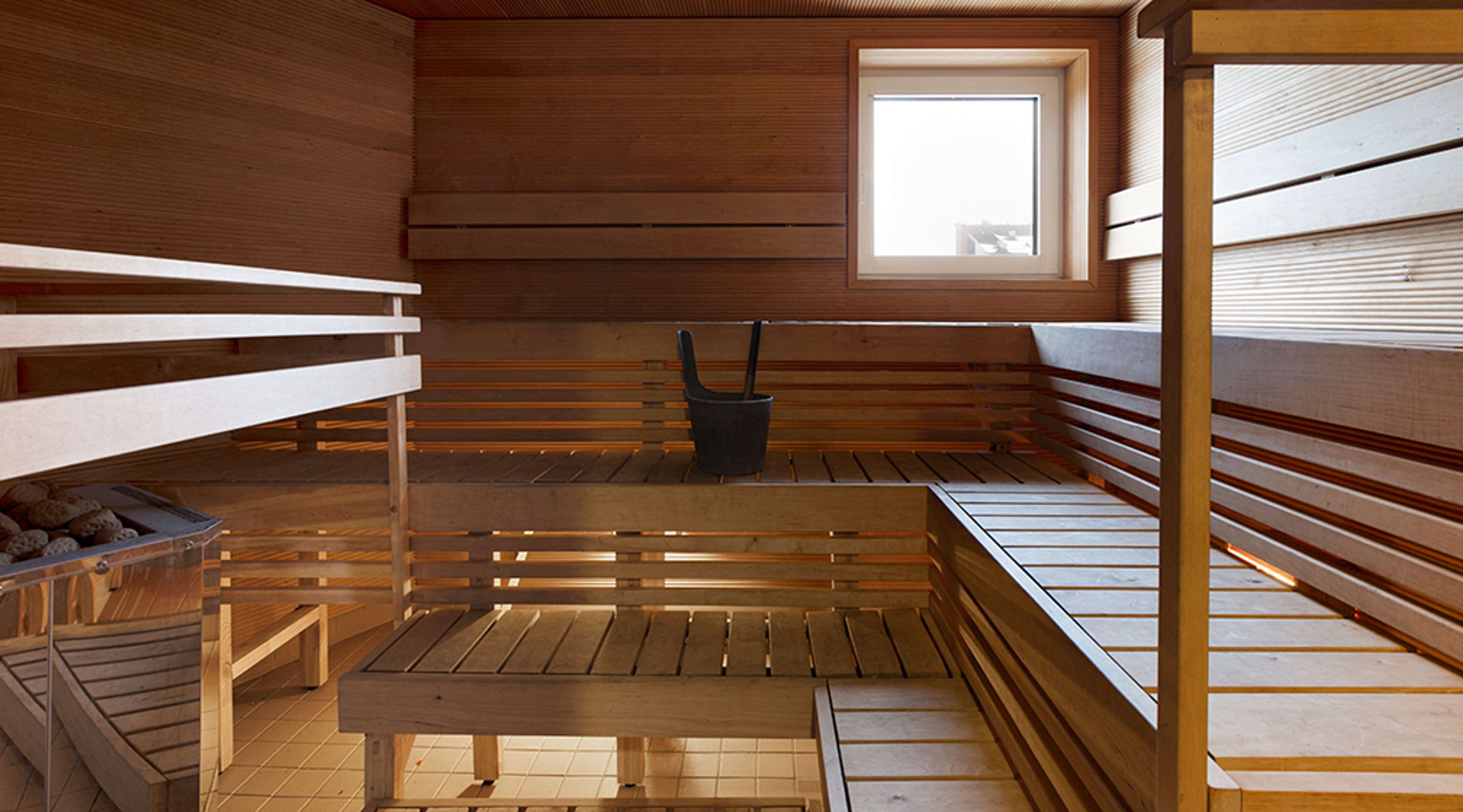 Moderni sauna puisilla lauteilla, joissa useita tasoja. Seinällä on pieni ikkuna ja lattialla löylykiulu.