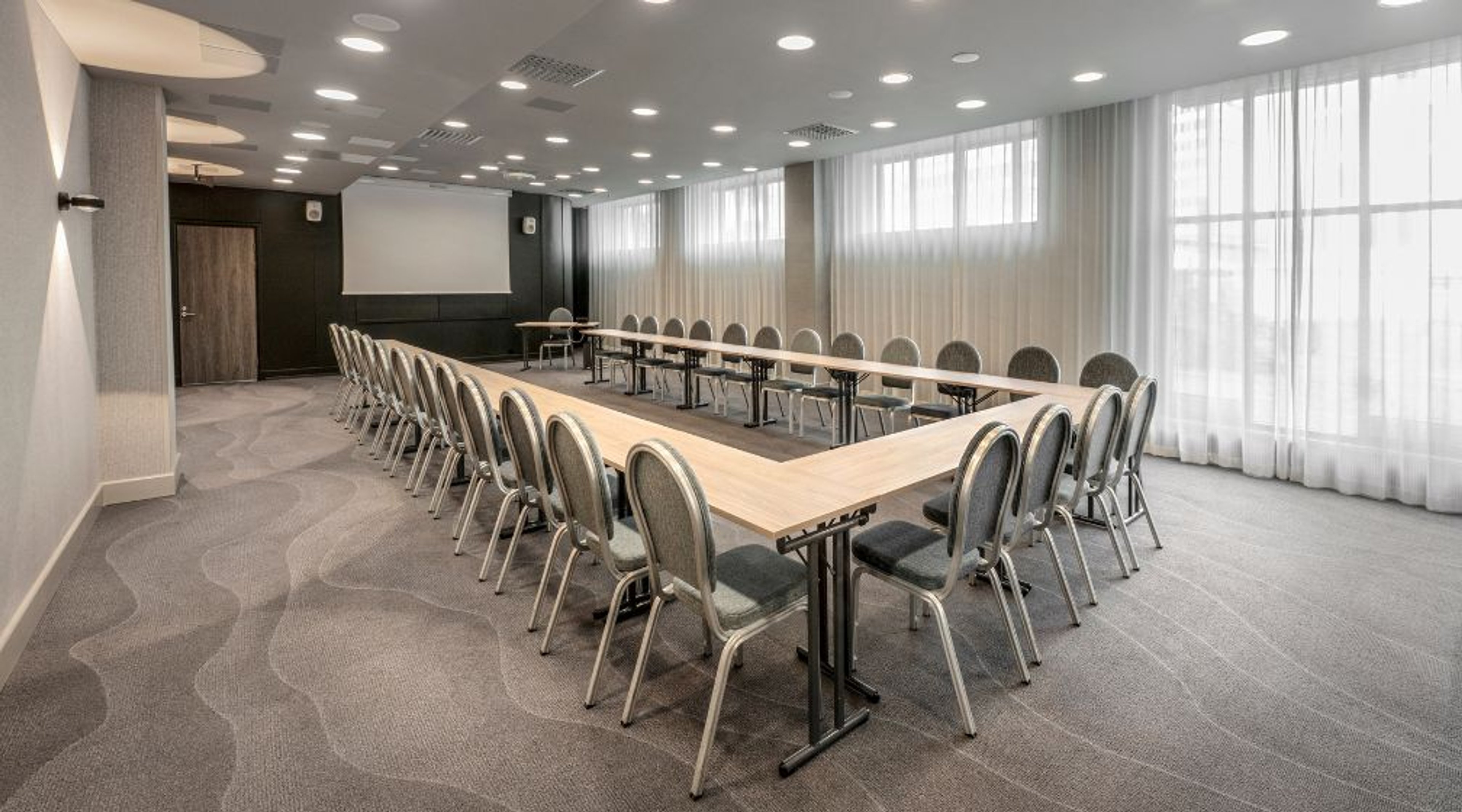 U-kujuline koosolekuruum heledavärviliste laudade ja toolidega, projektoriekraan, suured kardinad ja hea valgustus. Ühtlane vaipkate.