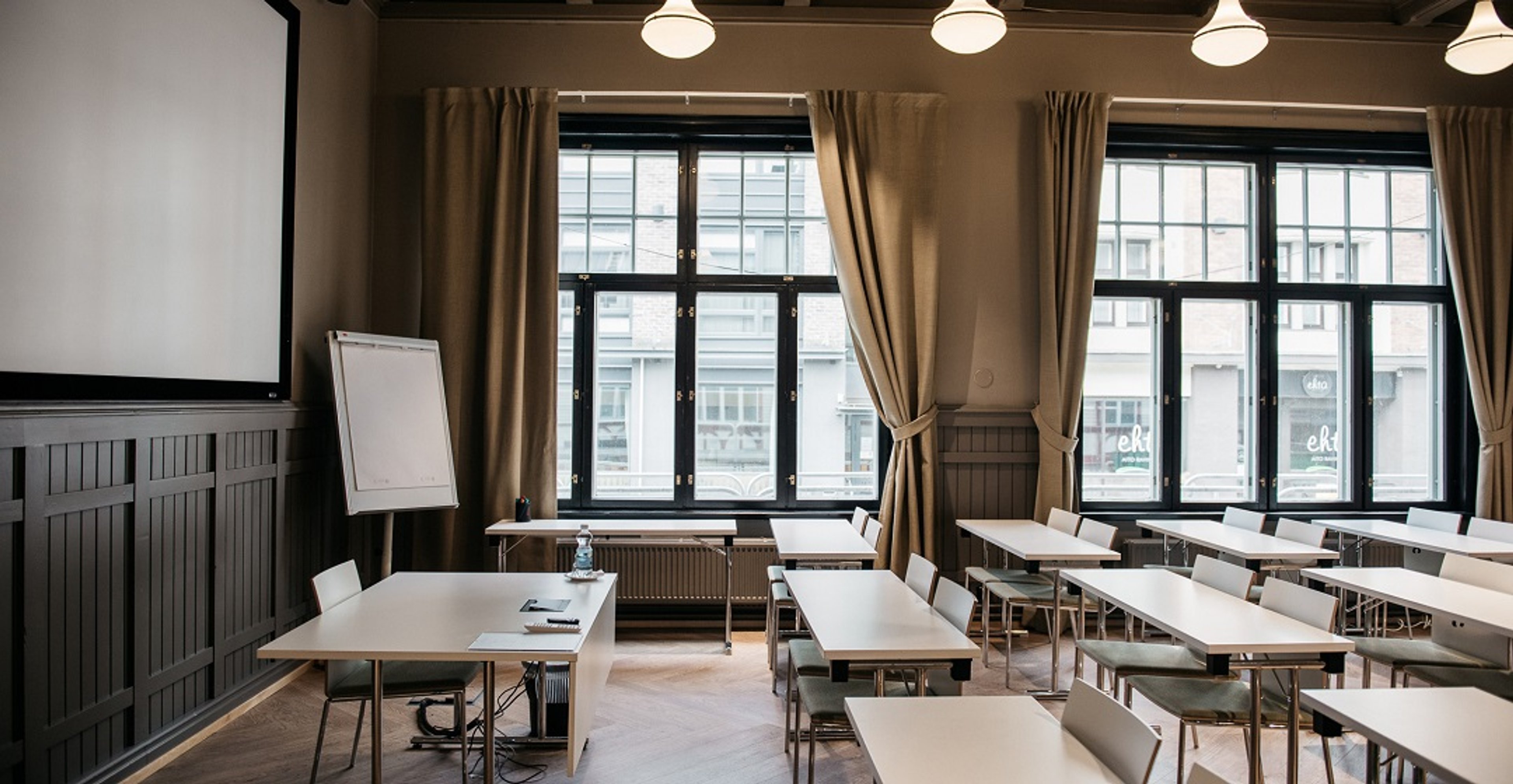 Kokoushuone, jossa valkoiset pöydät järjestetty riveihin, iso valkokangas edessä, suuret ikkunat beigen verhoin ja puinen lattia.