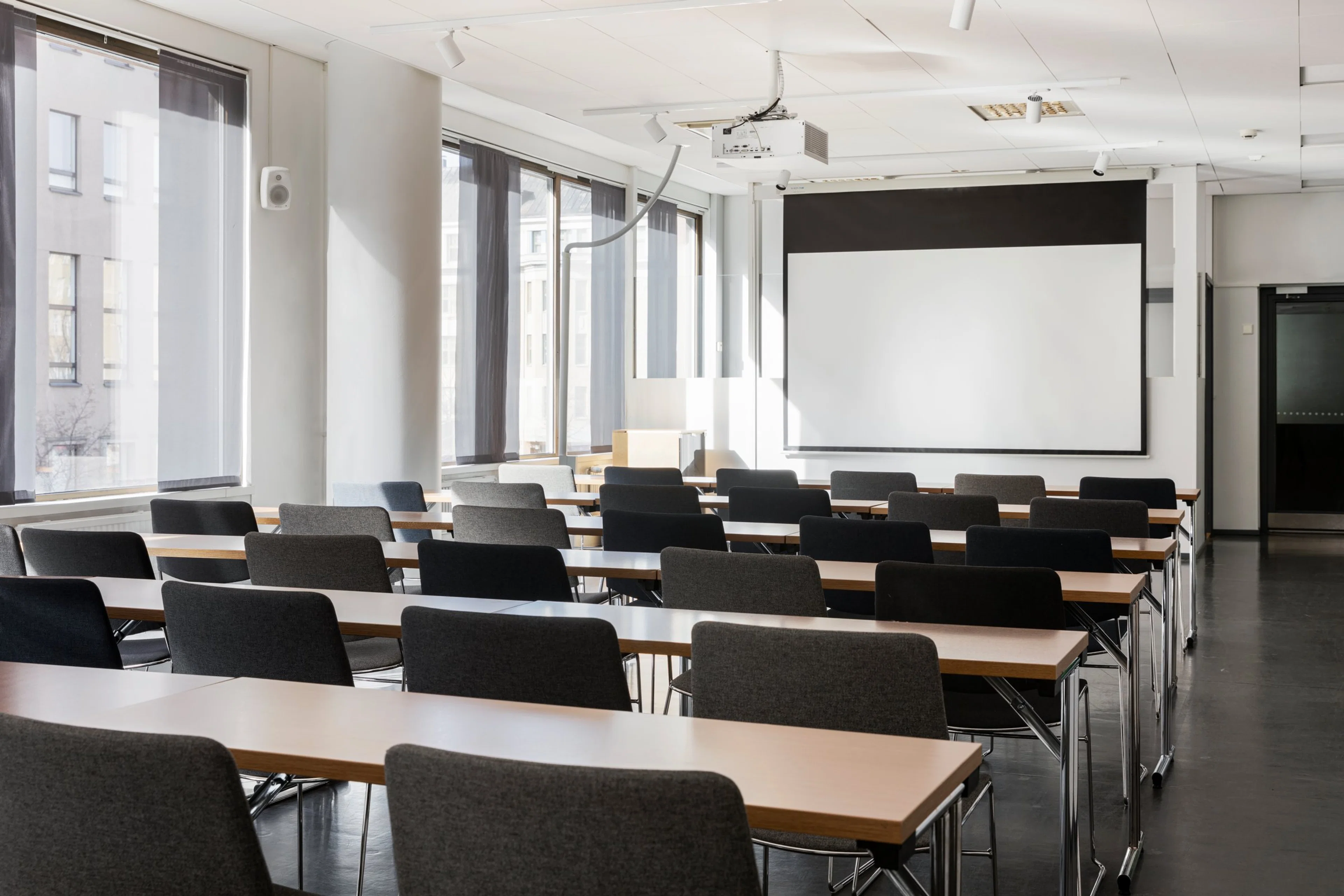 Valoisa kokoushuone, jossa on rivit pöytiä ja tuoleja. Edessä on valkokangas ja katossa projektori. Tilassa on suuret ikkunat.