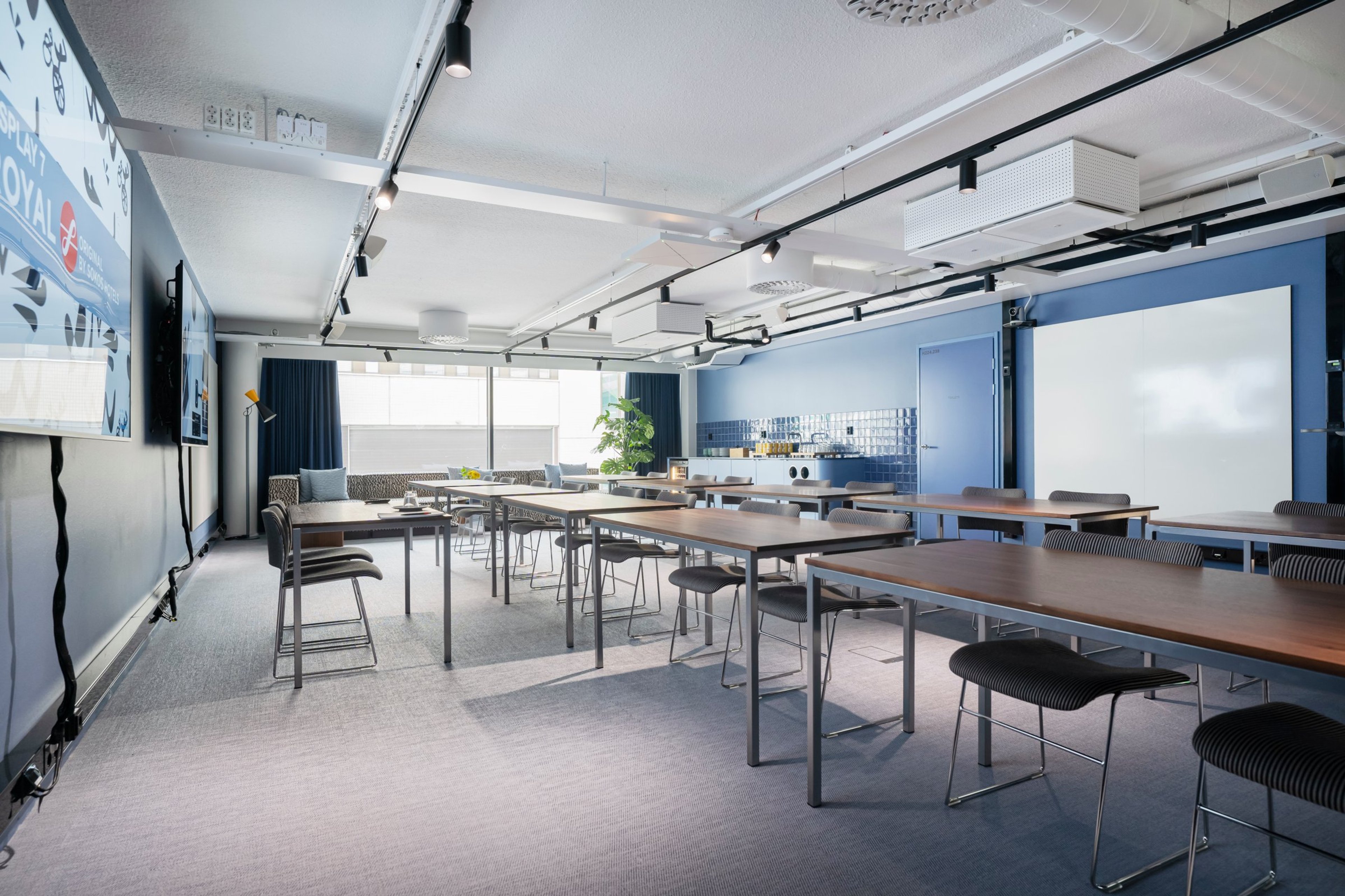 Moderni kokoushuone sinisillä seinillä, puupintaisilla pöydillä ja baarituoleilla. Huoneessa on raidallinen kokolattiamatto ja suuret ikkunat.