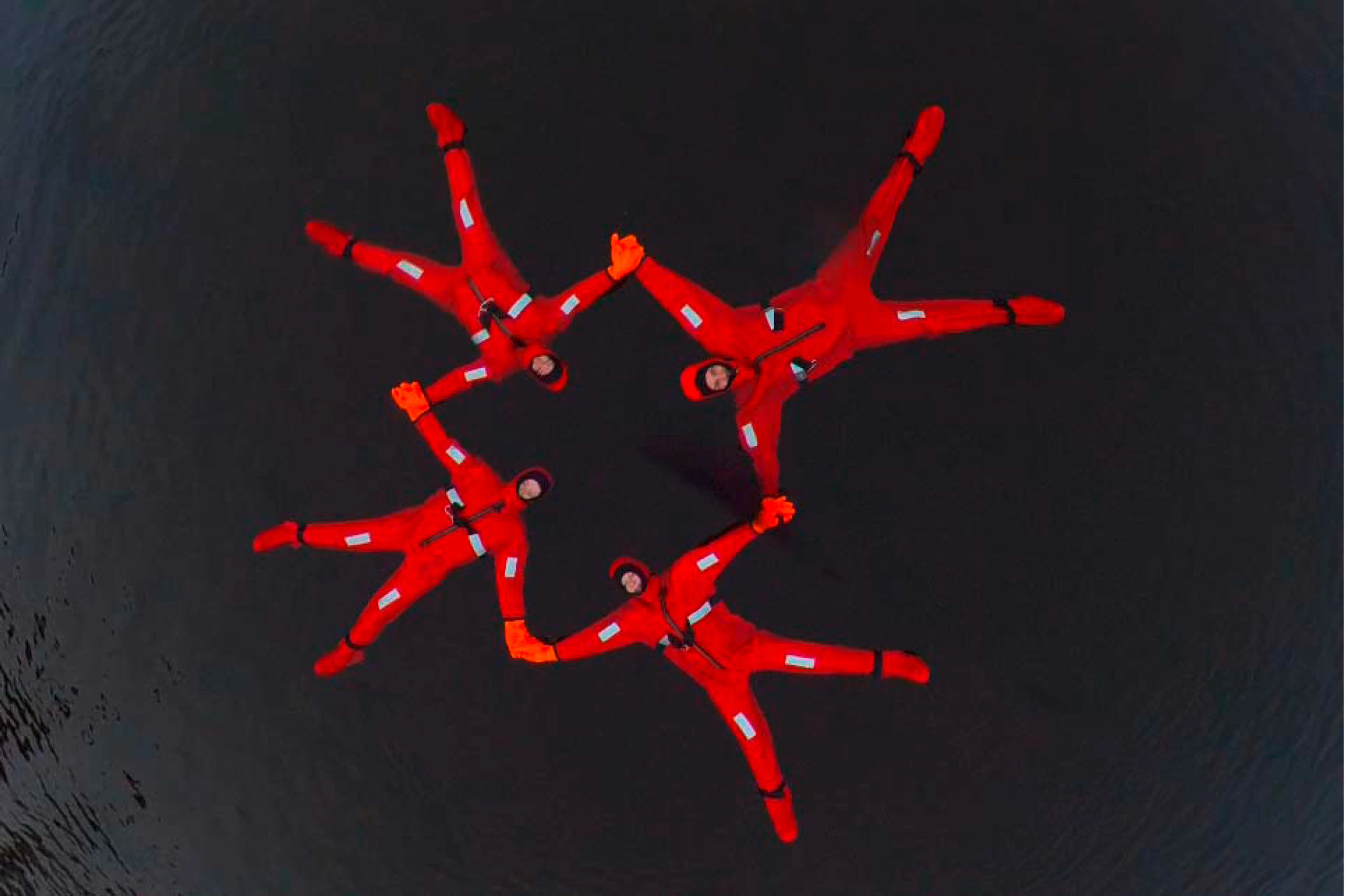 Aerial view of four people floating in dark water wearing bright red survival suits, forming a star-like formation.