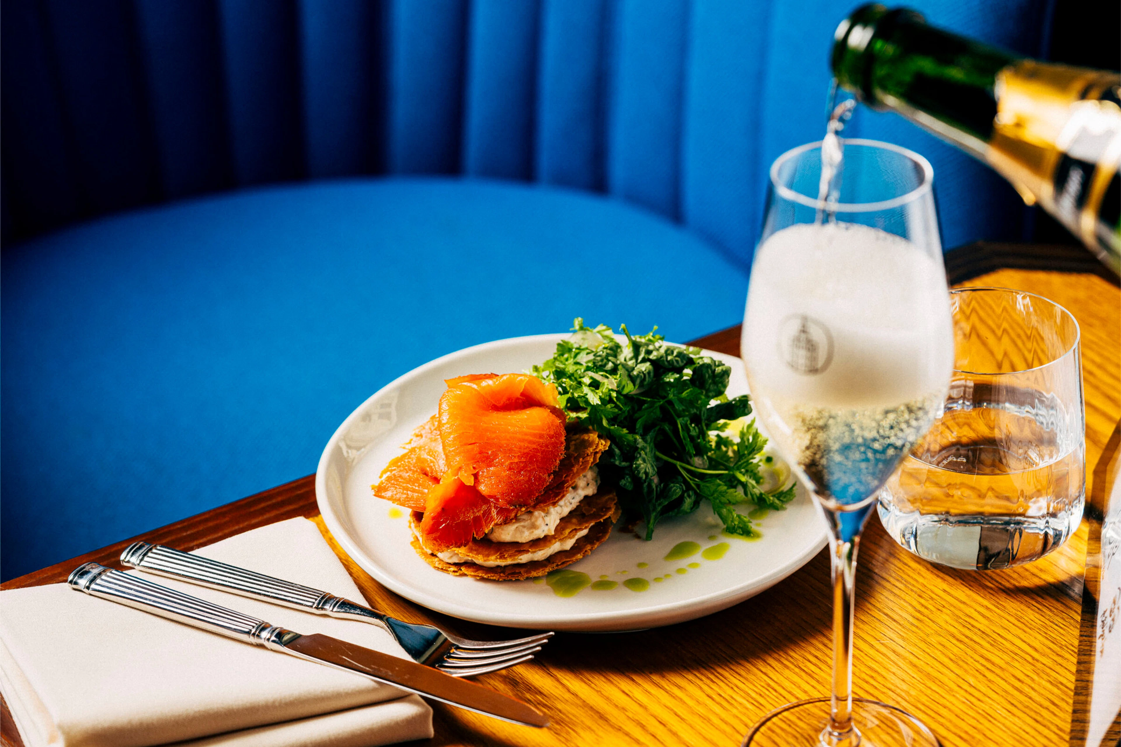 Dish with smoked salmon and green herbs on a white plate. Next to it, champagne being poured into a flute and a water glass on a wooden table.