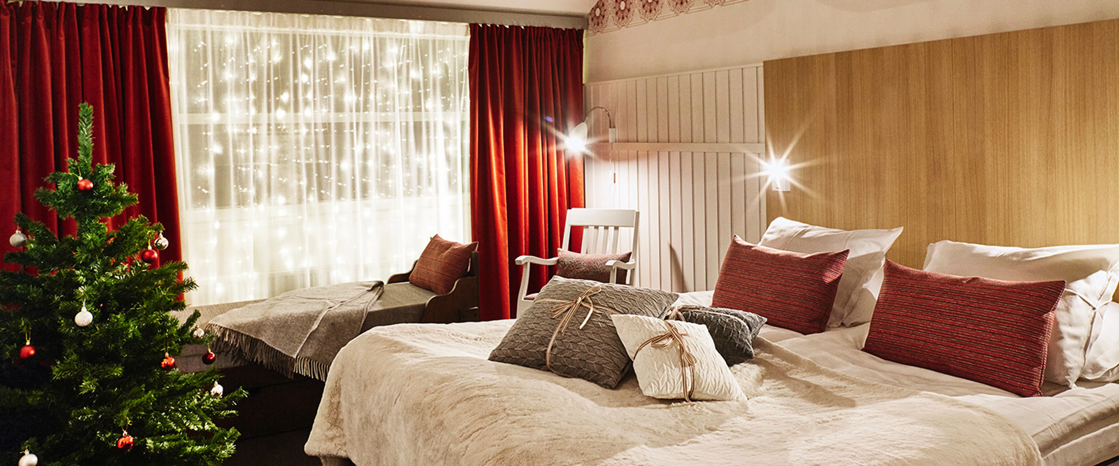 Cozy hotel room with large bed, lounge chair, red curtains and Christmas lights on the window. A small decorated Christmas tree stands in the corner.