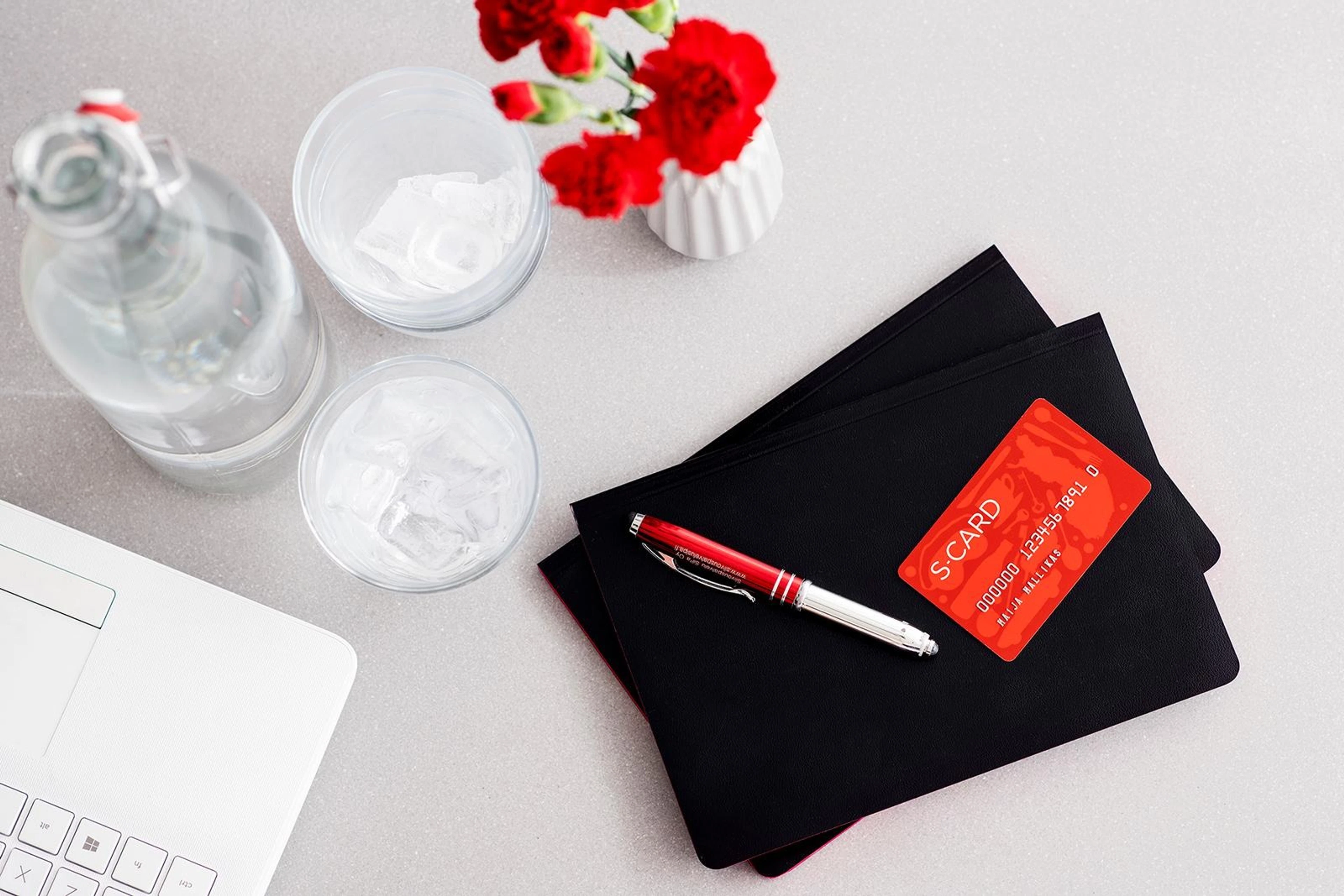 Desk with laptop, water bottle, glasses, flower vase and black notebooks with pen.