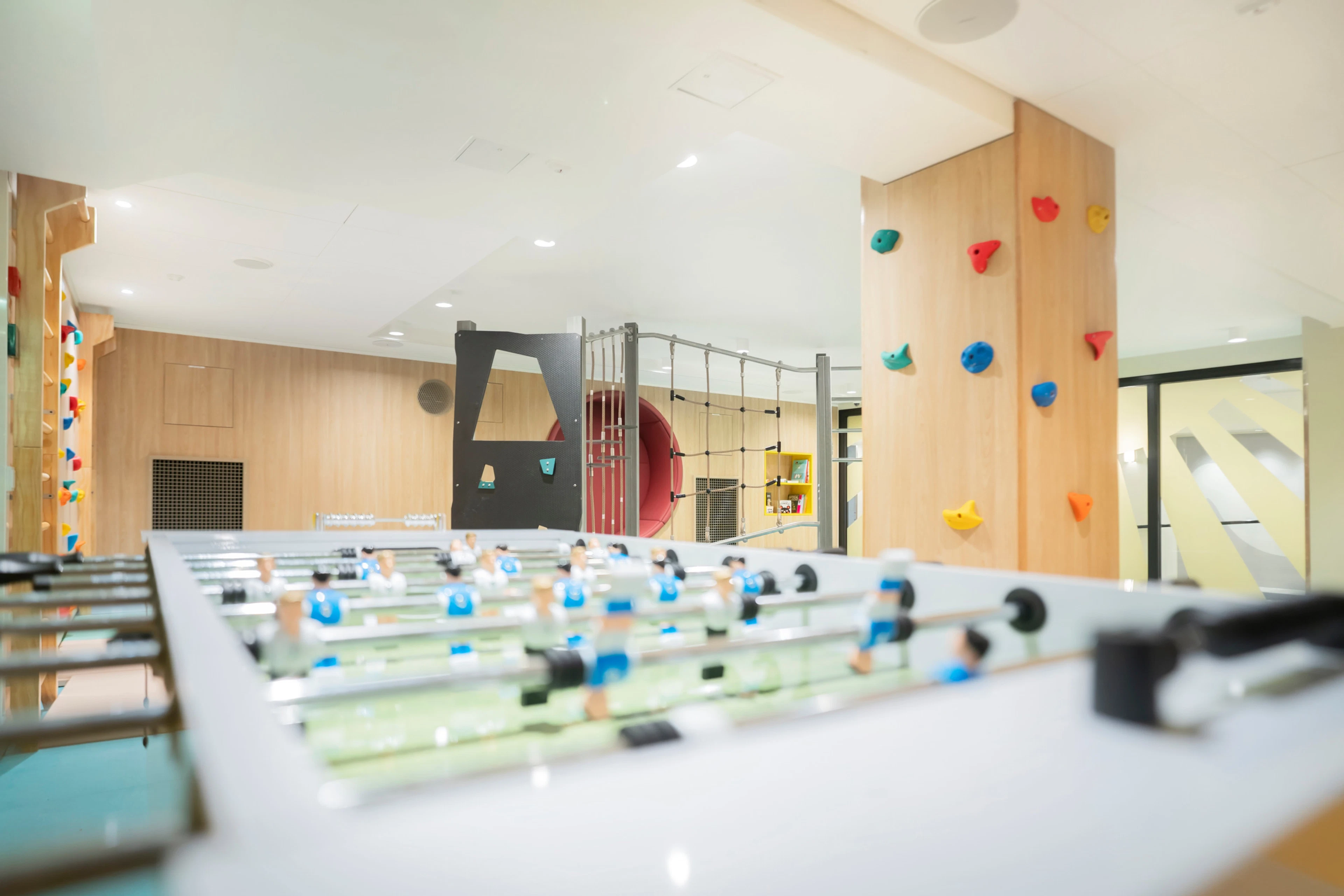 Valoisa leikkihuone, jossa on pöytäjalkapallo, kiipeilyseinä värikkäillä otteilla ja monipuolinen kiipeilyteline taustalla.