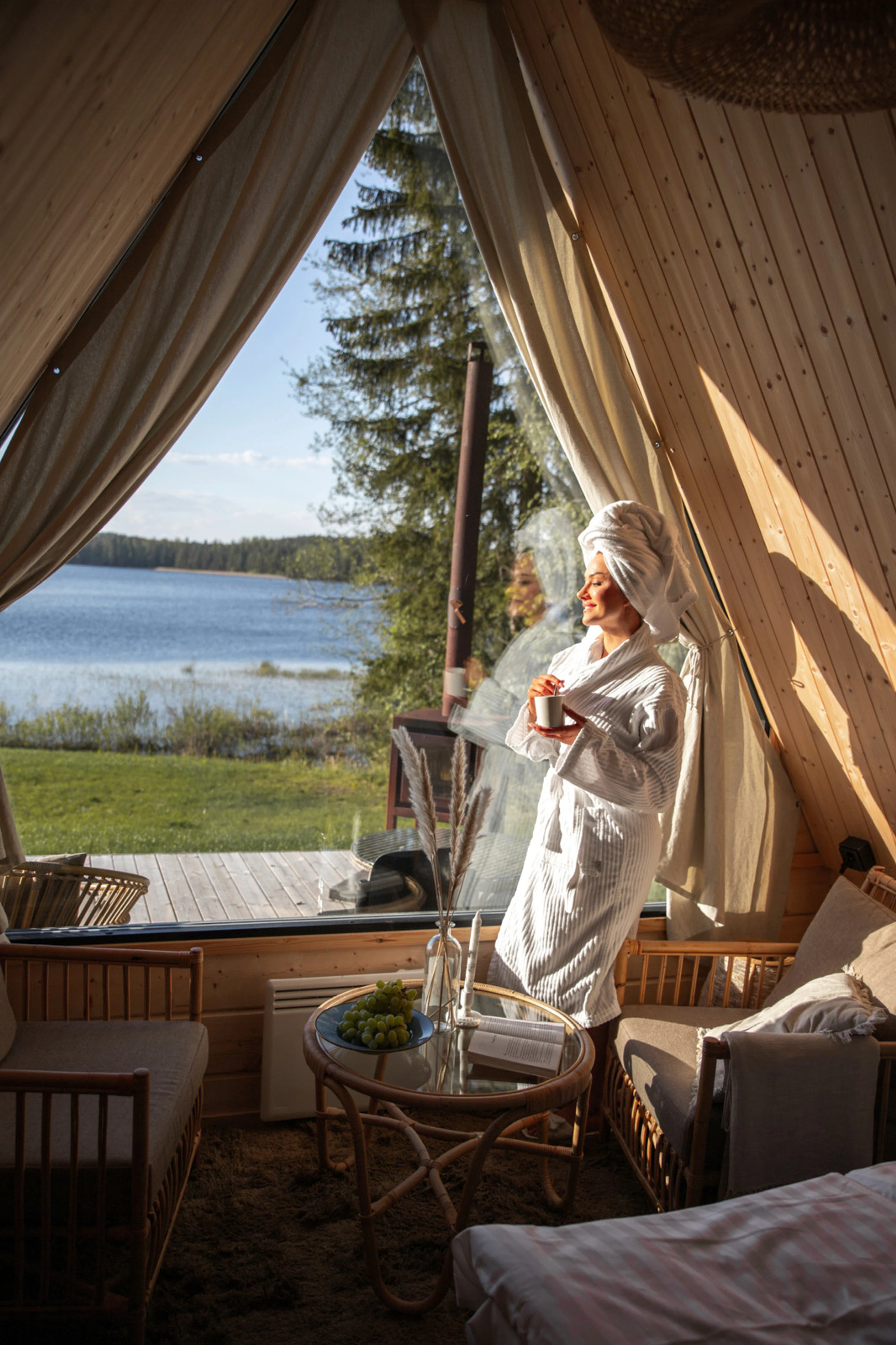 Hymyilevä nainen sekoittaa kahvia ja katsoo aurinkoiselle järvelle laawun ikkunasta.