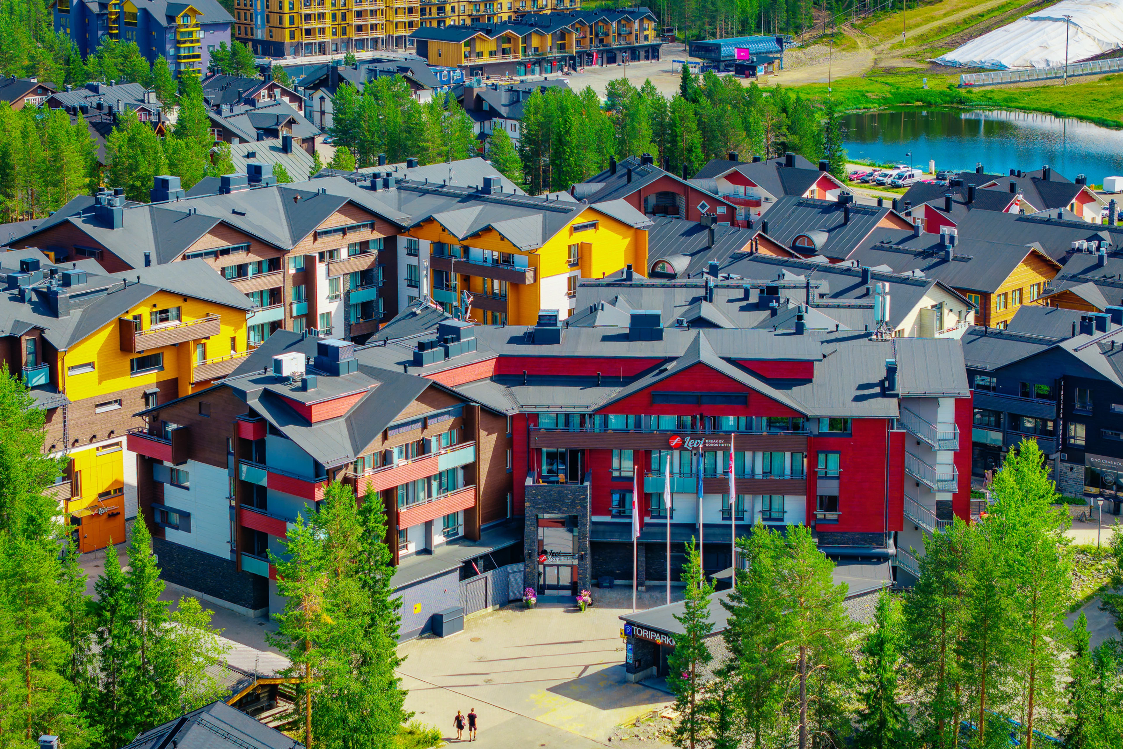 Flygfoto över semesterort, färgglada bostadsbyggnader omgivna av tallar och platt asfaltgata.