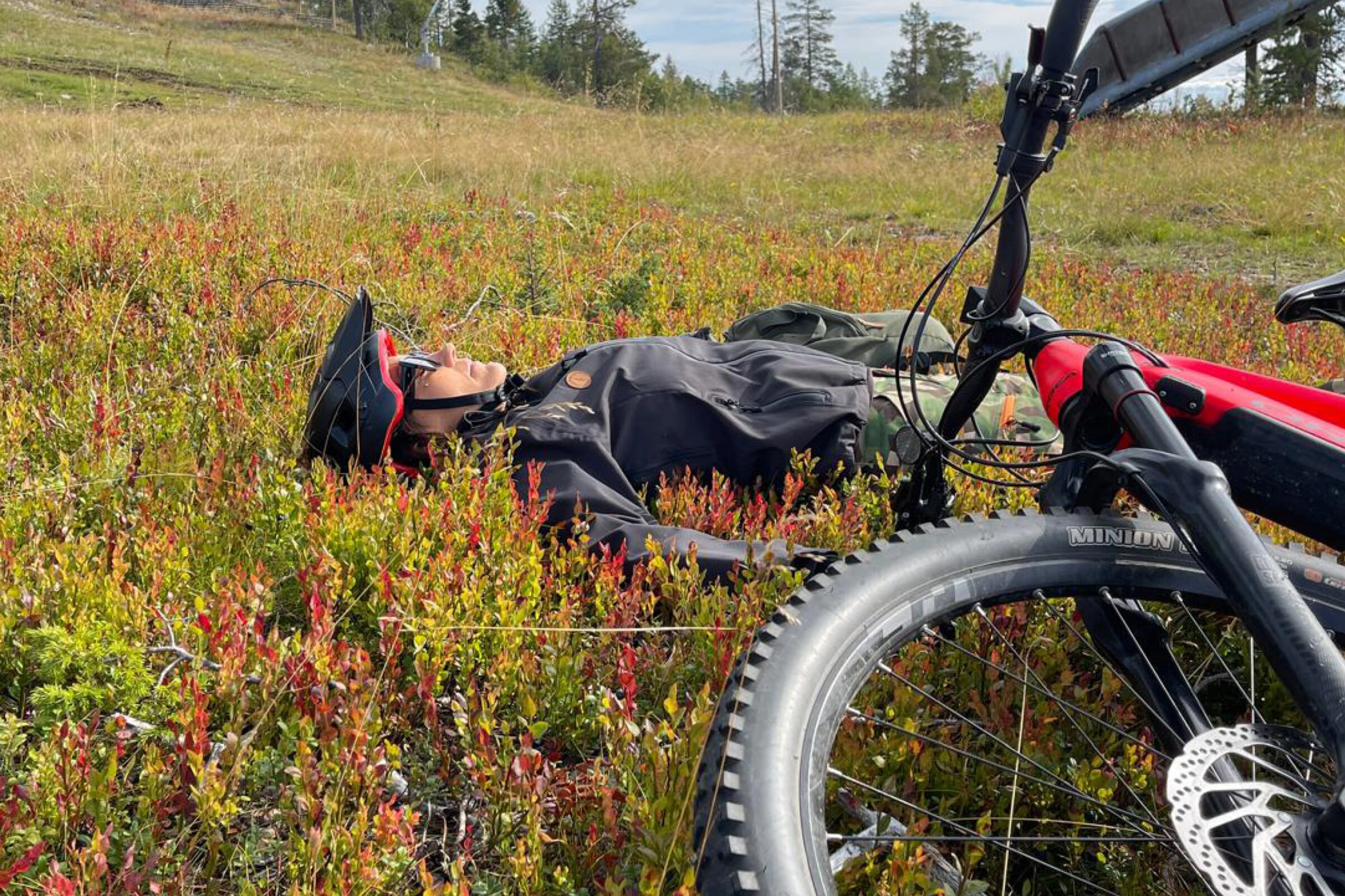 Cyklist vilar bland färgglad höstvegetation, cykel ligger på gräsmattan bredvid. Ojämn terräng med låg vegetation.