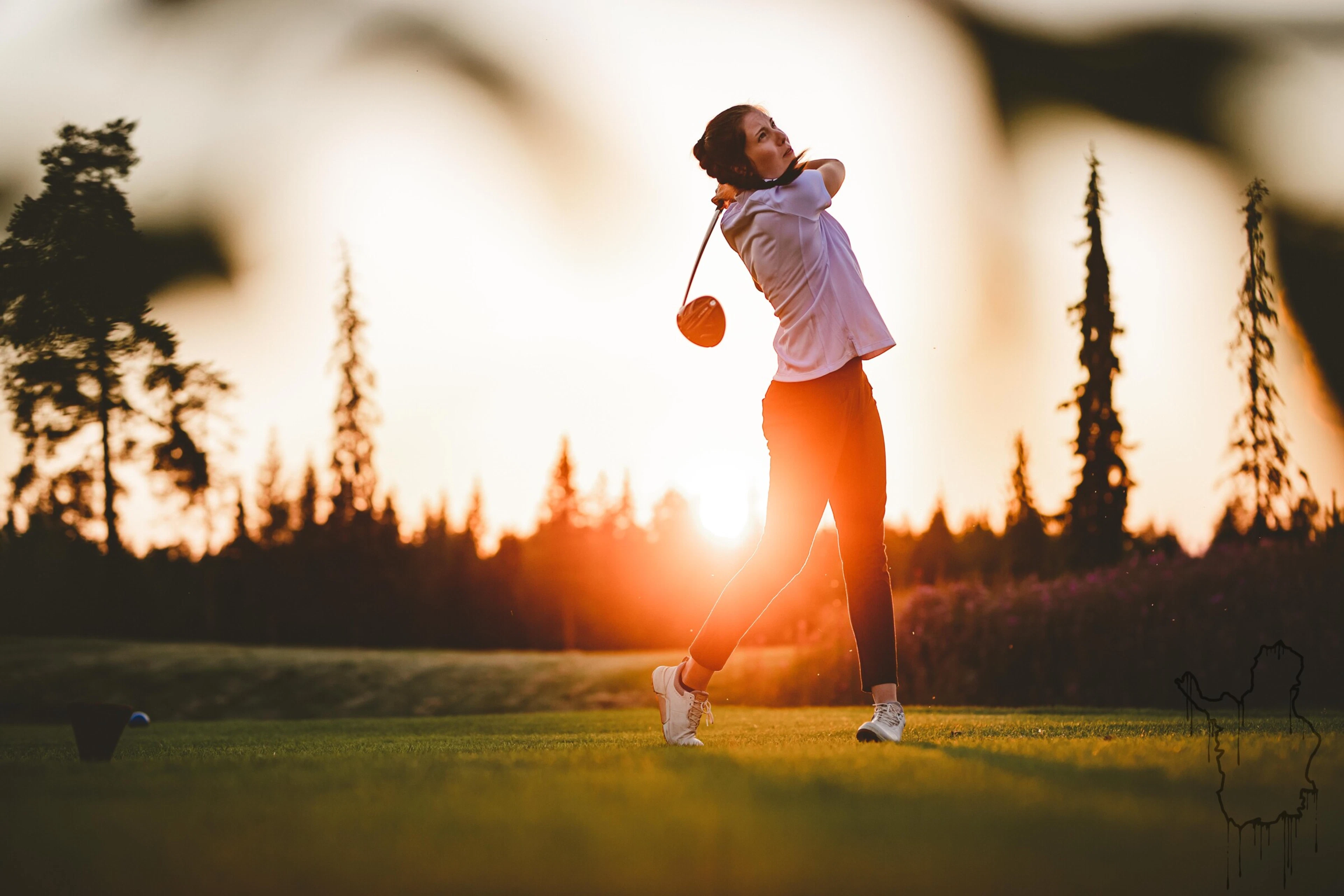 Golfer swinging on a level grass surface at sunset with forest in background.