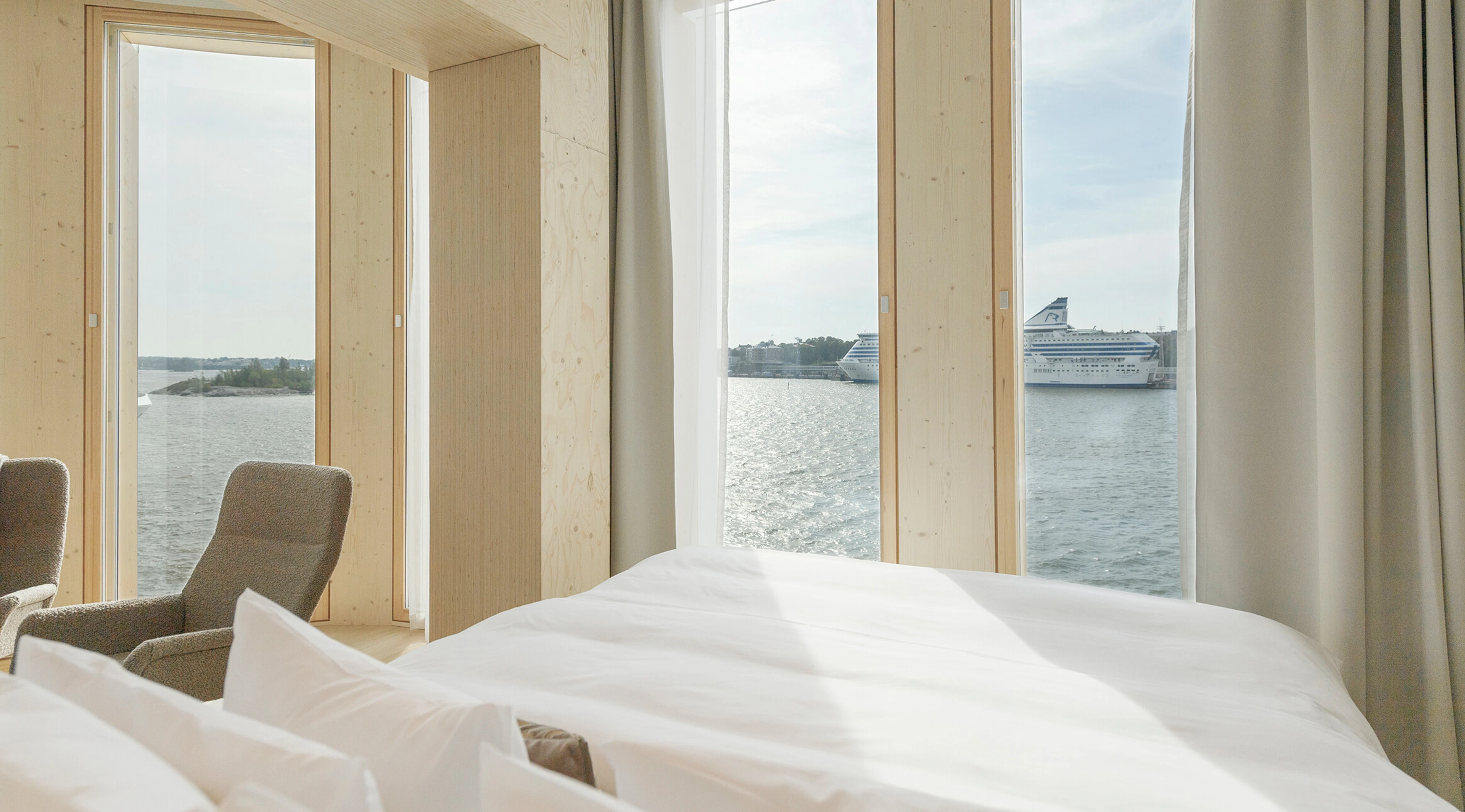 Hotel room with large window overlooking the sea and light wood paneling on walls.