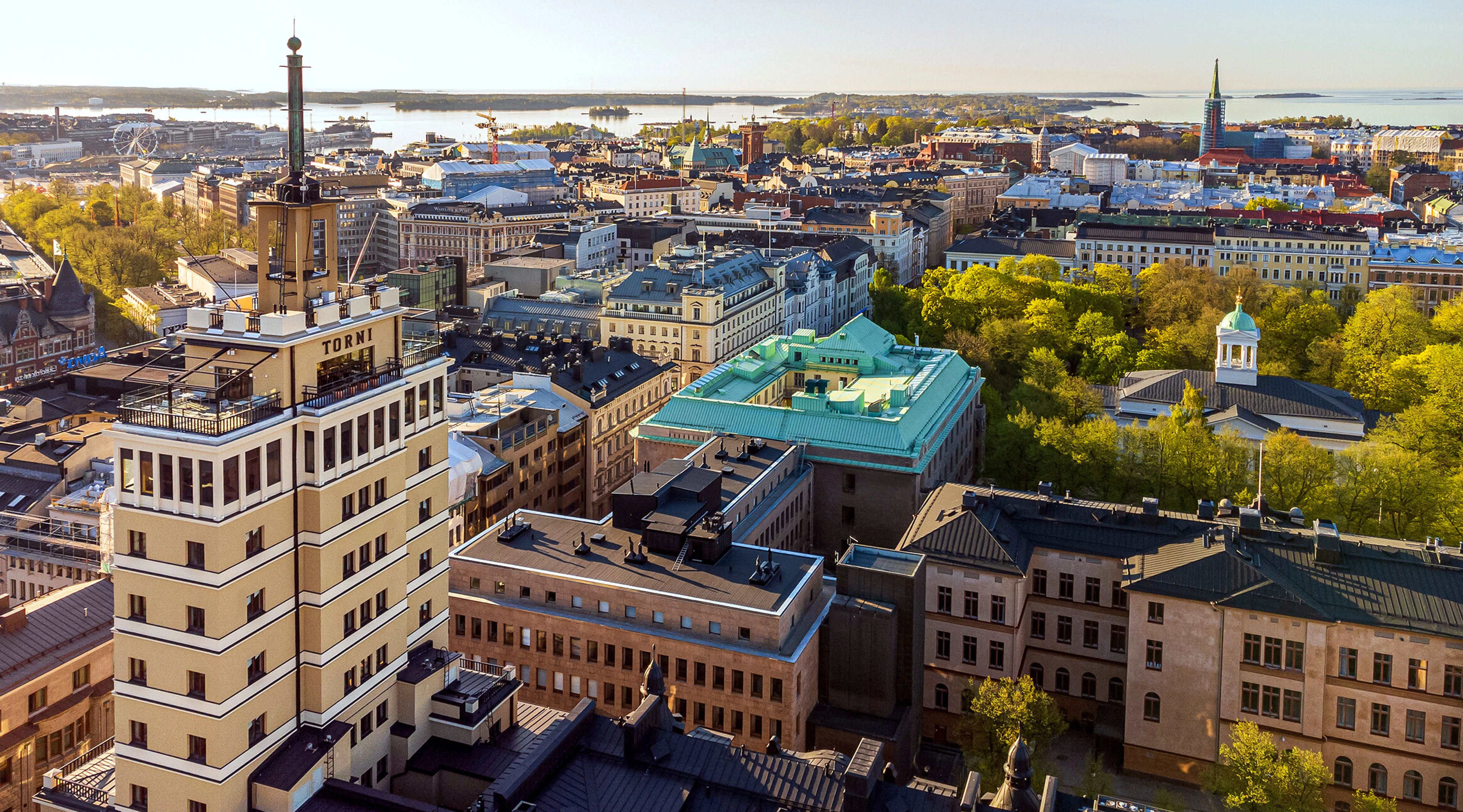Aerial view of Helsinki city center. Buildings with green roofs stand out among the rooftops. A bay and islands visible in the background.