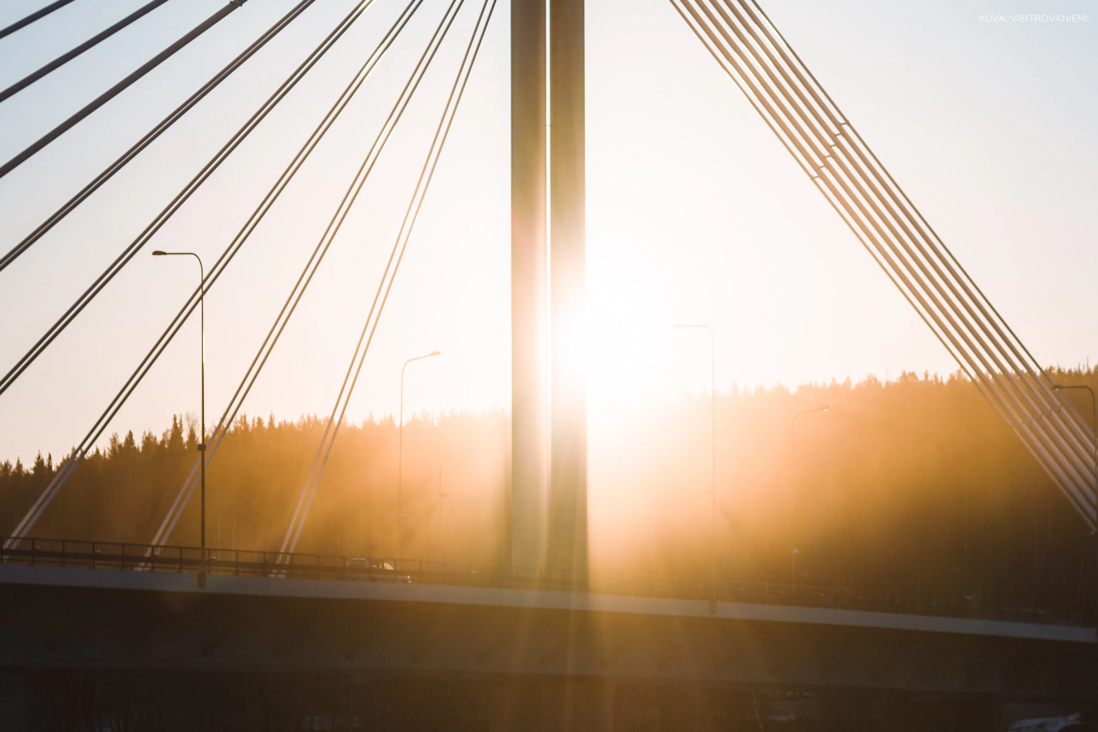 Silta auringonlaskussa, siluetin muodostavat kaapelit ja tukipylväs, taustalla metsäinen horisontti kultaisessa valossa.