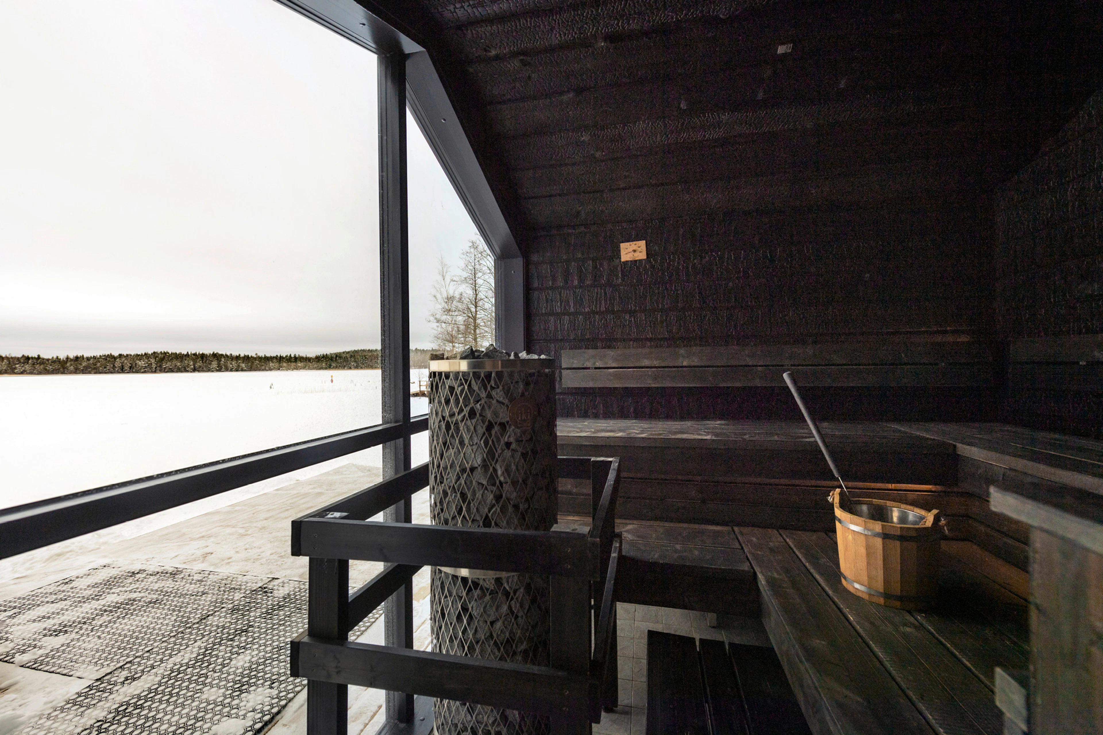 Tumma puusauna laajalla ikkunalla, josta näkyy luminen järvimaisema.