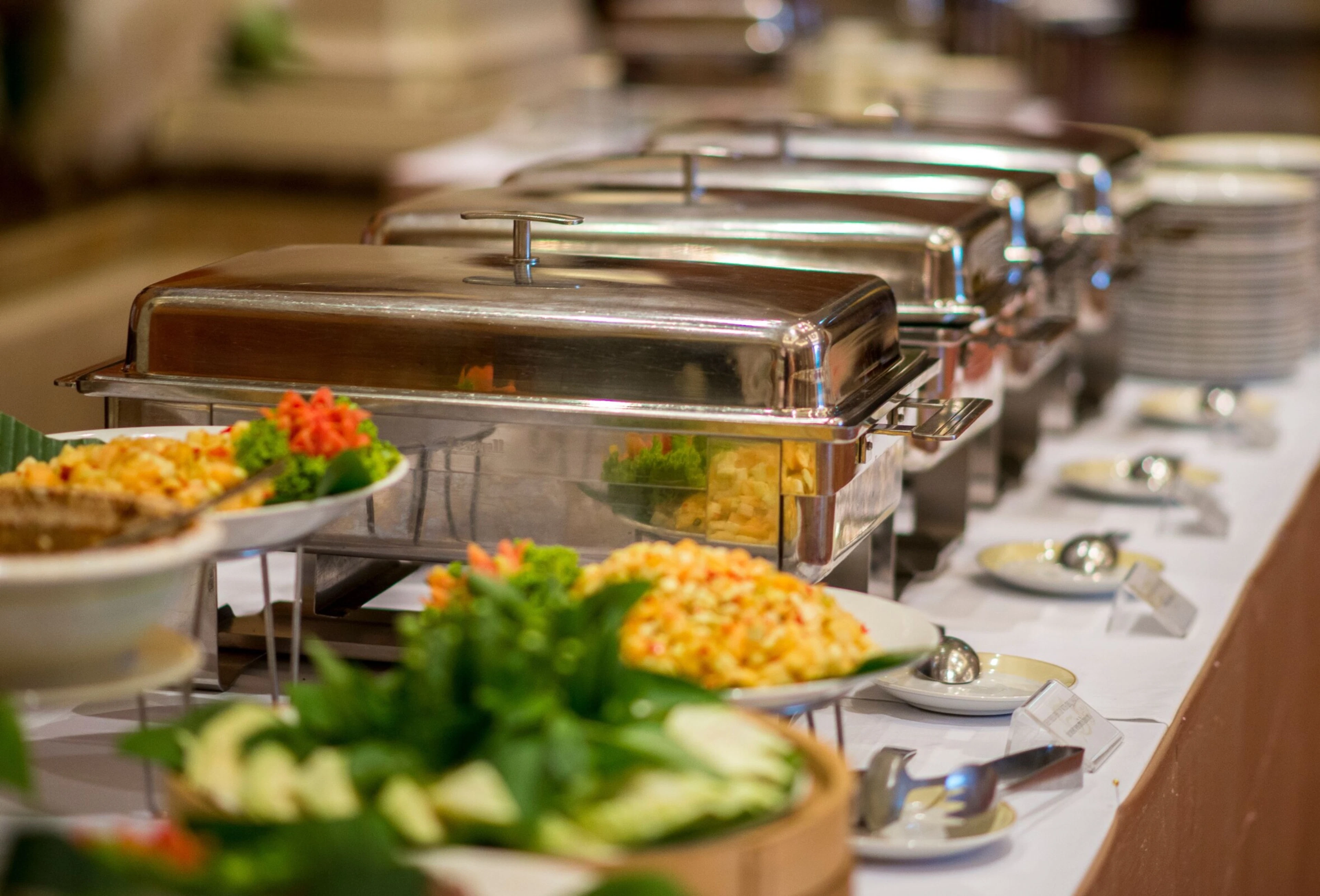 Buffet table with multiple chafing dishes with metal lids and plates with salads.