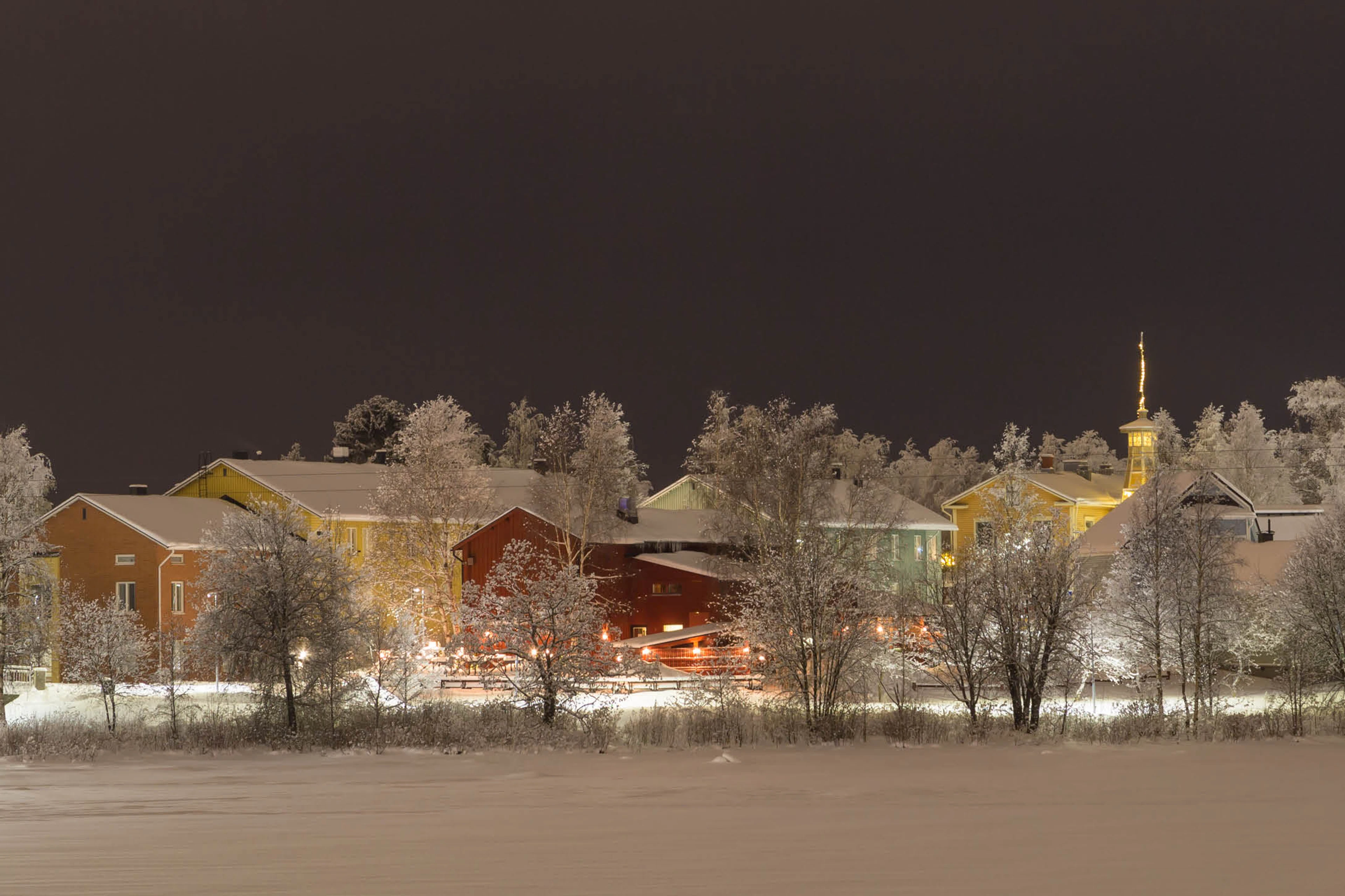 Talvinen yömaisema lumisesta pikkukaupungista. Värikkäitä taloja ja kirkontorni hohtavat valaistuksessa, puut ovat huurteen peitossa.