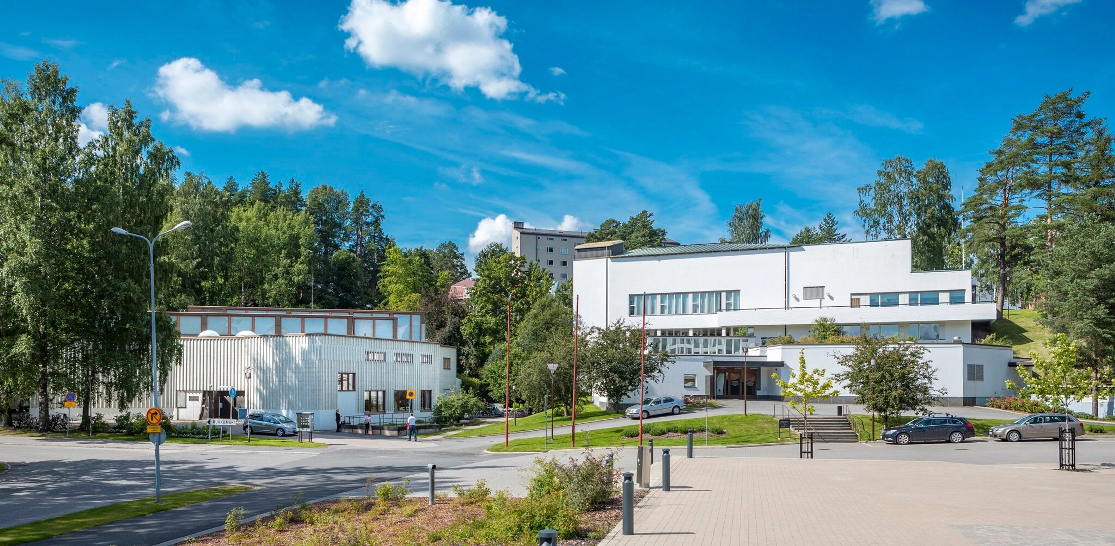 Moderni valkoinen rakennuskompleksi ympäröivine viherkaistoineen. Rakennuksen edustalla asfaltoidut tiet ja pysäköintialue, portaat sisäänkäynnille.