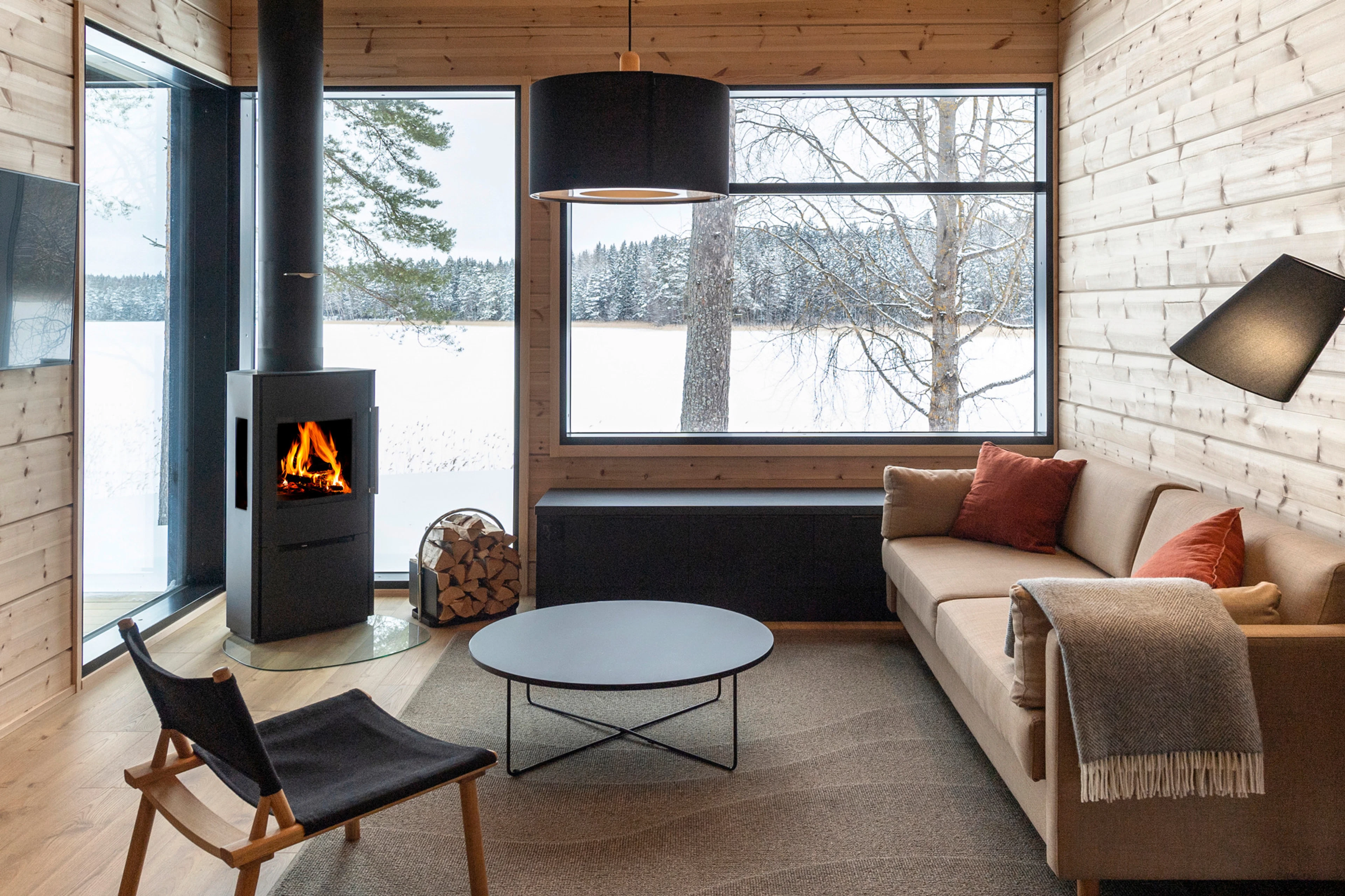 Moderni hirsihuvila puusisustuksella. Takka, sohva ja suuret ikkunat talviseen järvimaisemaan.