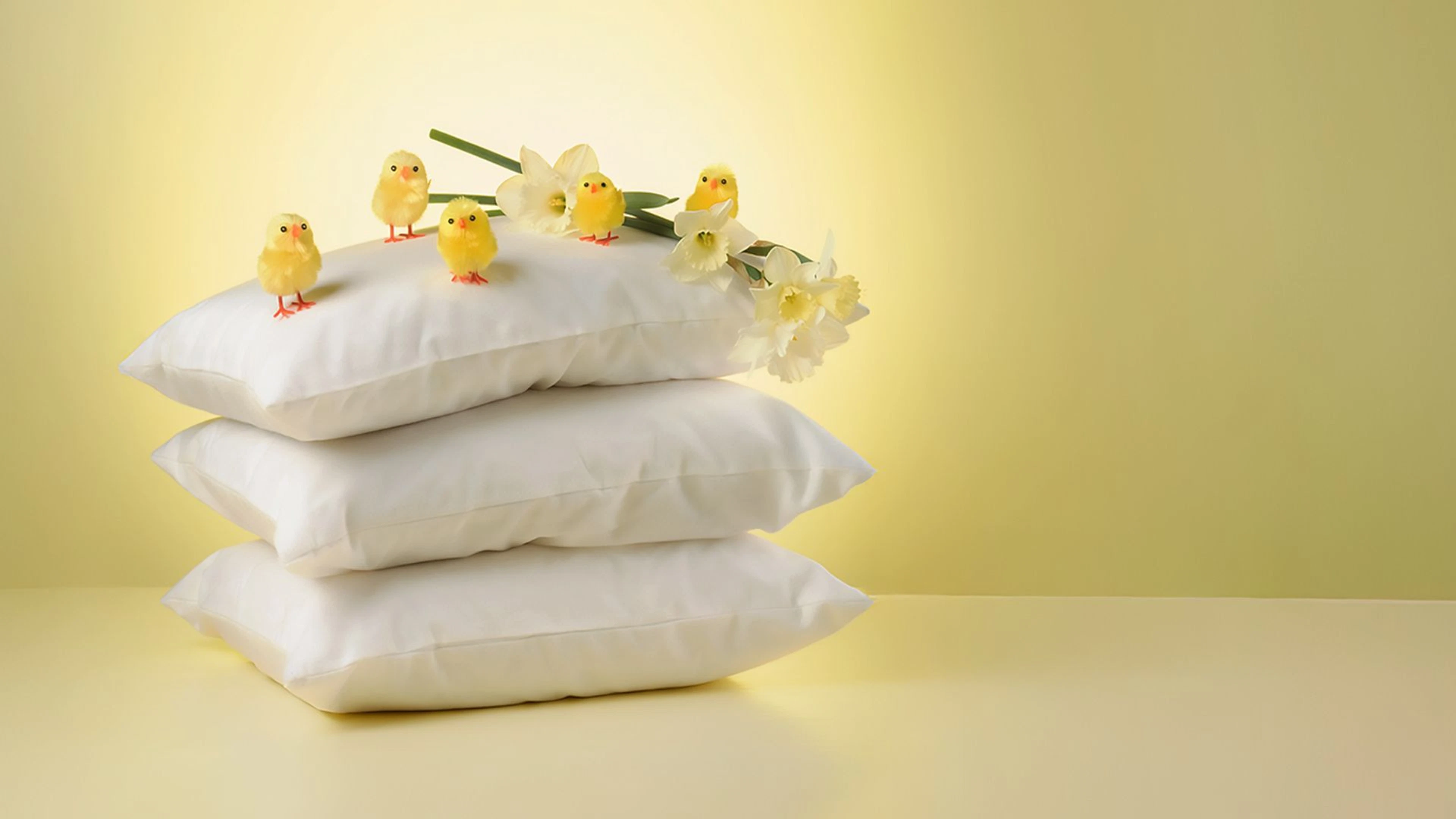 Kolme valkoista tyynyä pinossa, päällimmäisellä viisi pientä keltaista pääsiäistipua ja valkoisia narsisseja keltaisella taustalla.