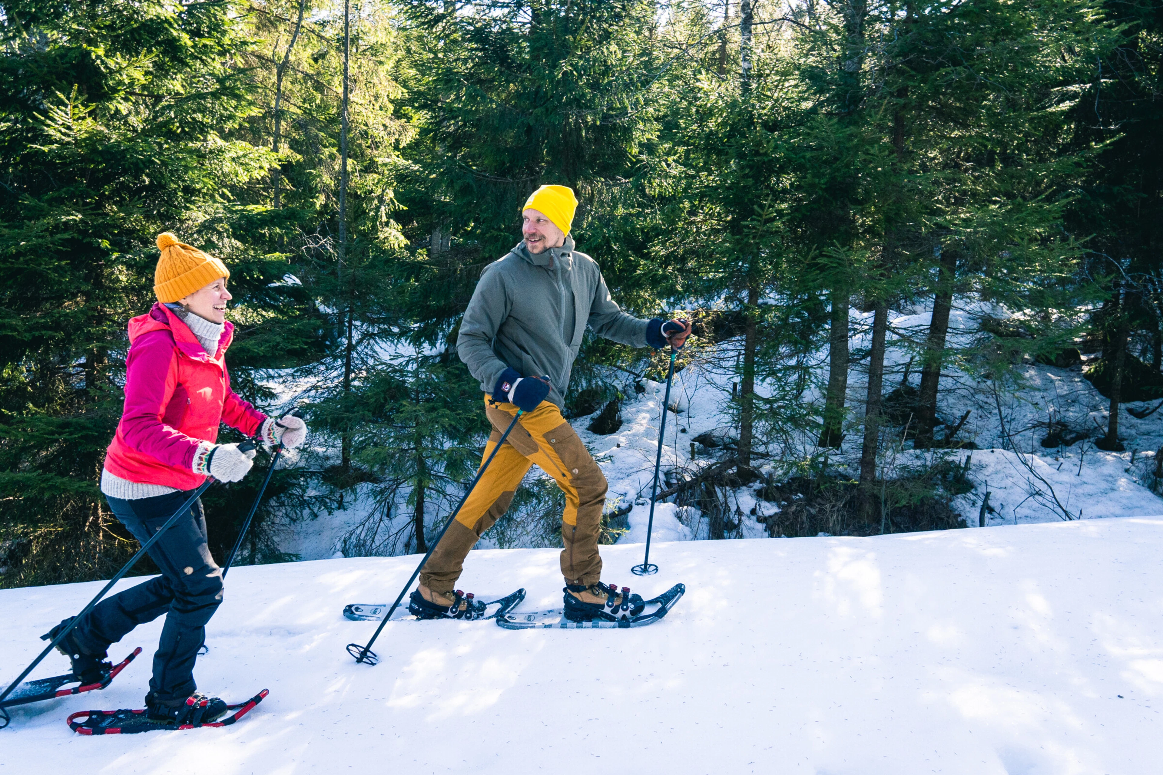 Kaksi ihmistä lumikenkäilemässä talvisessa metsässä, kirkkaat pipot ja talvivarusteet yllään.
