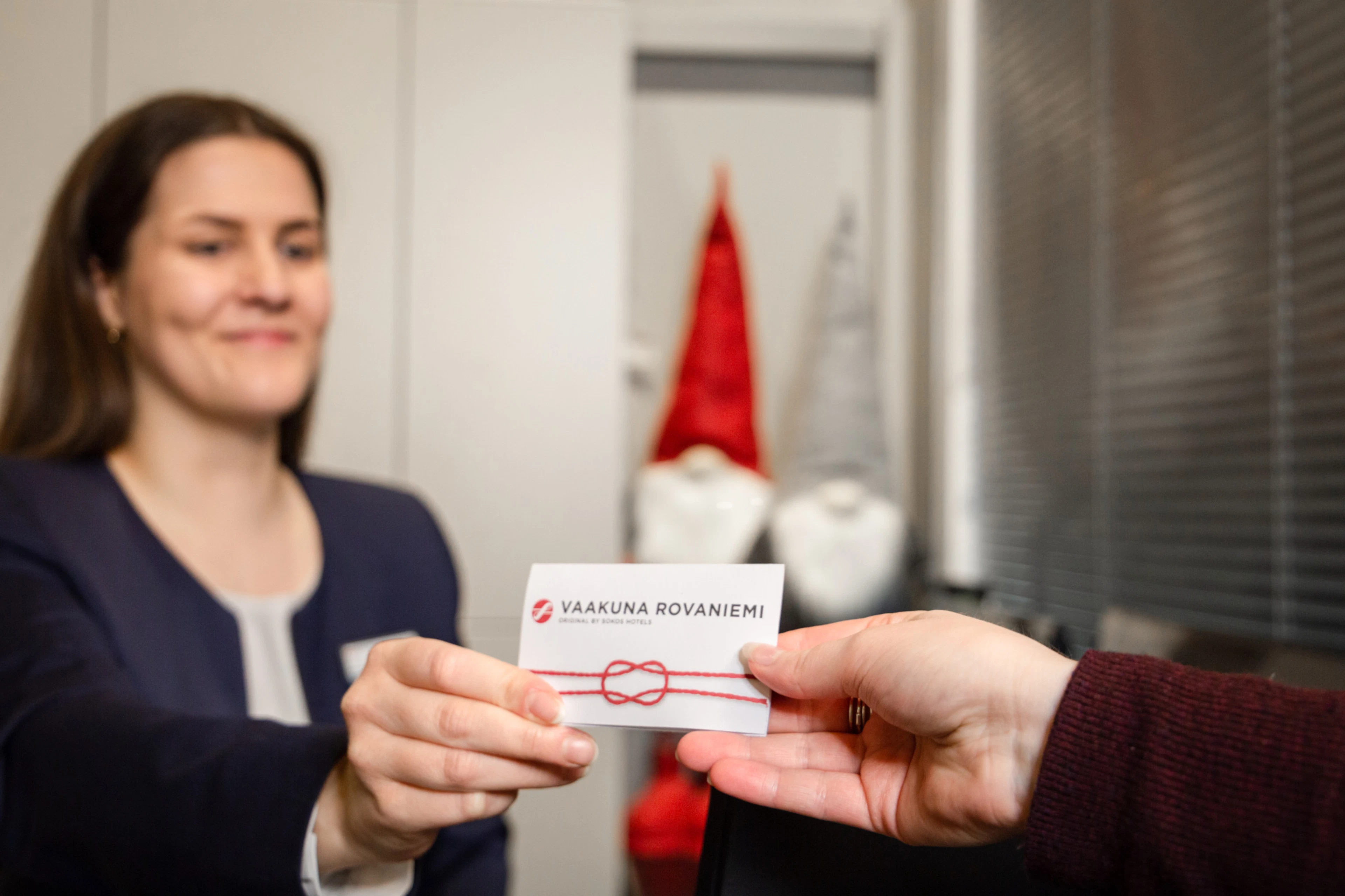 Person in a dark blue suit handing a Vaakuna Rovaniemi hotel card to another person. A Christmas gnome is visible in the background.